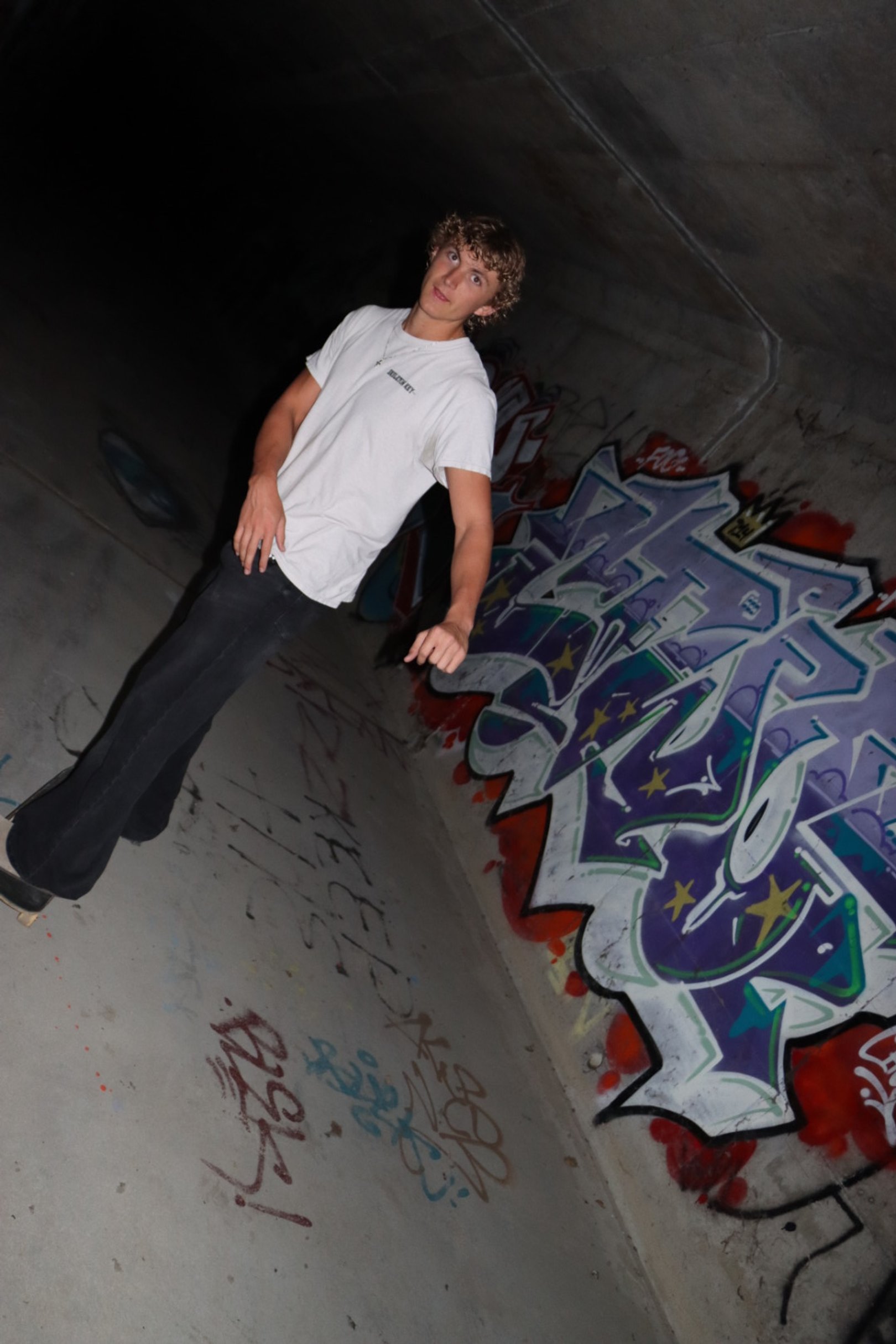 A young man with curly hair in a white t-shirt and black pants standing in an underground skate park with graffiti on the walls.