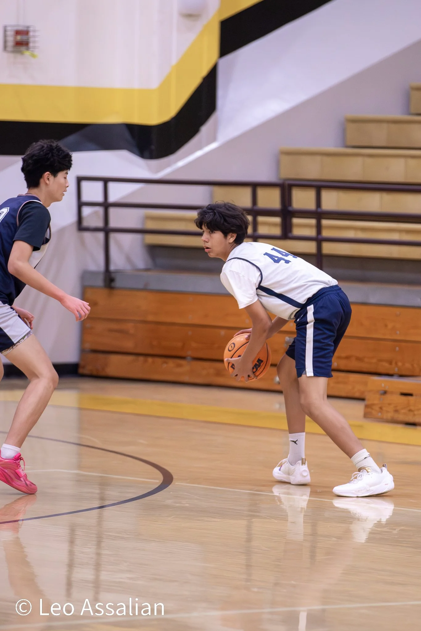 Two boys playing basketball in a gymnasium, one is dribbling the ball while the other is defending.