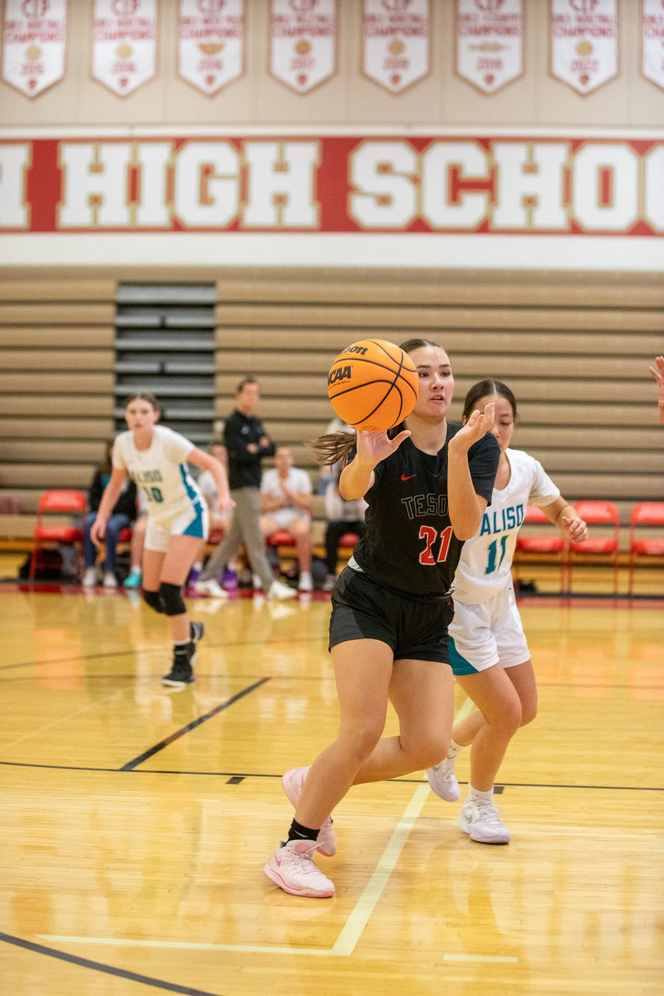 A young female basketball player in black uniform dribbling a basketball on an indoor gym court with other players and spectators in the background.