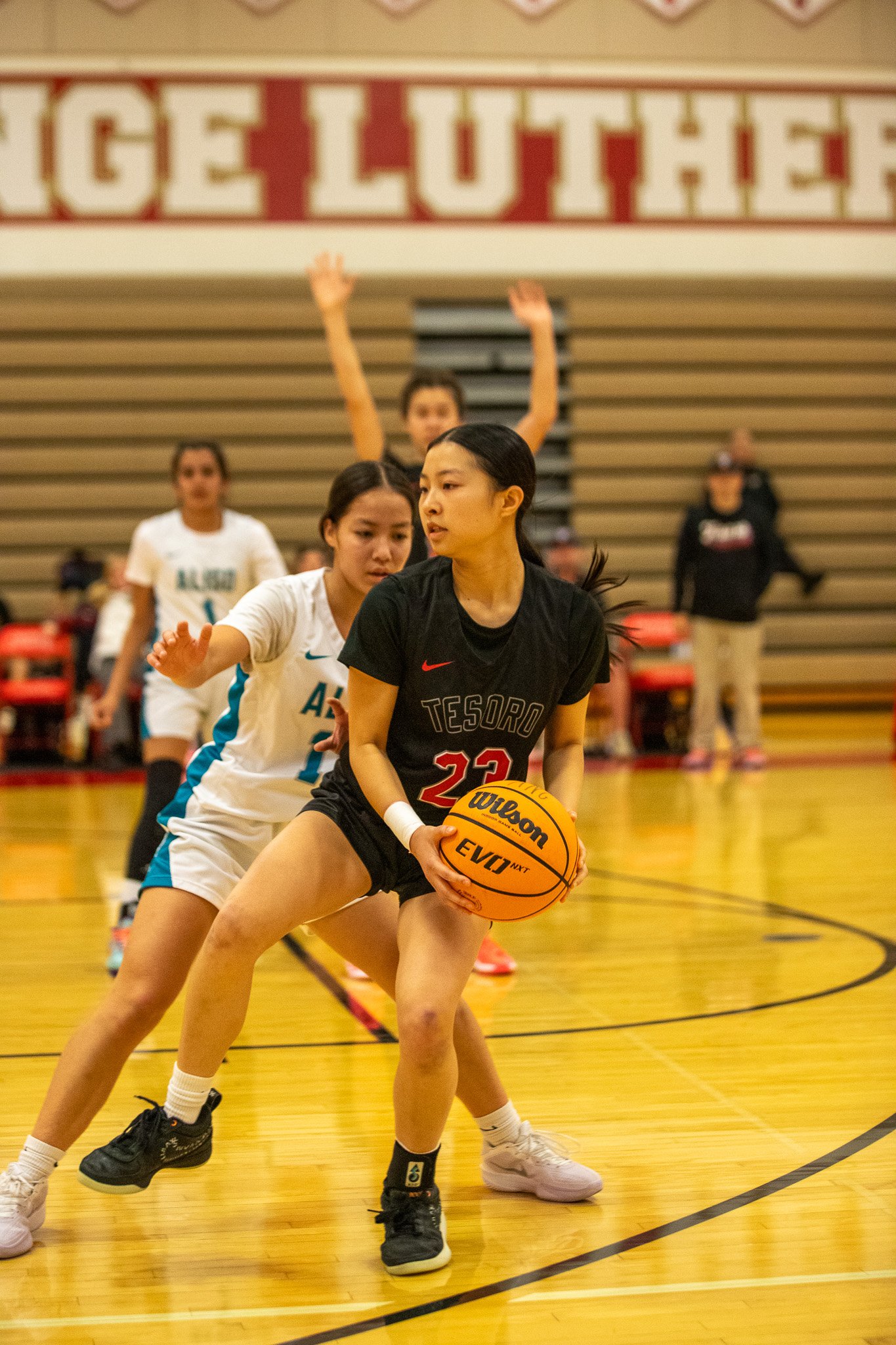Young female basketball player in black jersey holding a basketball, guarded closely by a female opponent in a white jersey, in an indoor gymnasium during a game.