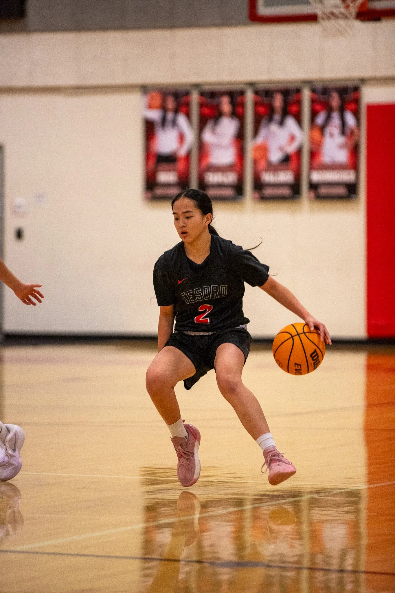 A young female basketball player in black uniform with the number 2 practicing dribbling on an indoor basketball court.