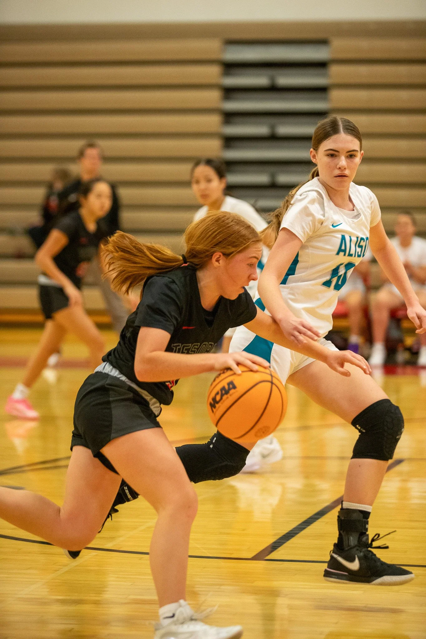 Girls playing basketball during a game, with one girl in black driving with the ball and another girl in white defending, on a gymnasium floor.