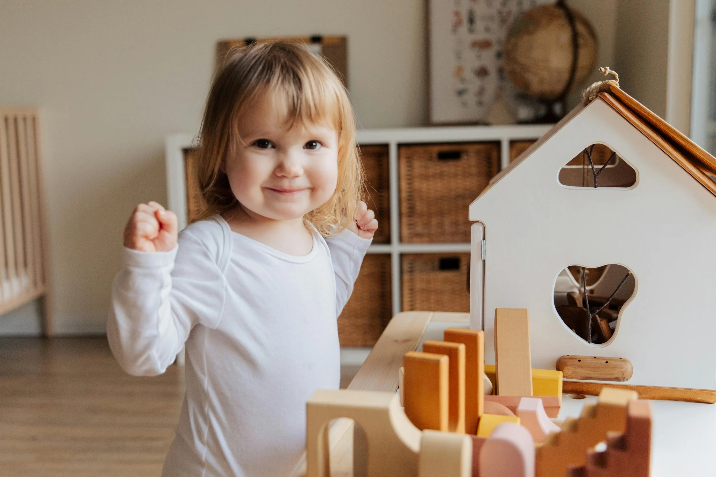 A young girl with blonde hair smiling and raising her fists in front of a wooden toy house and colorful blocks on a white table indoors.
