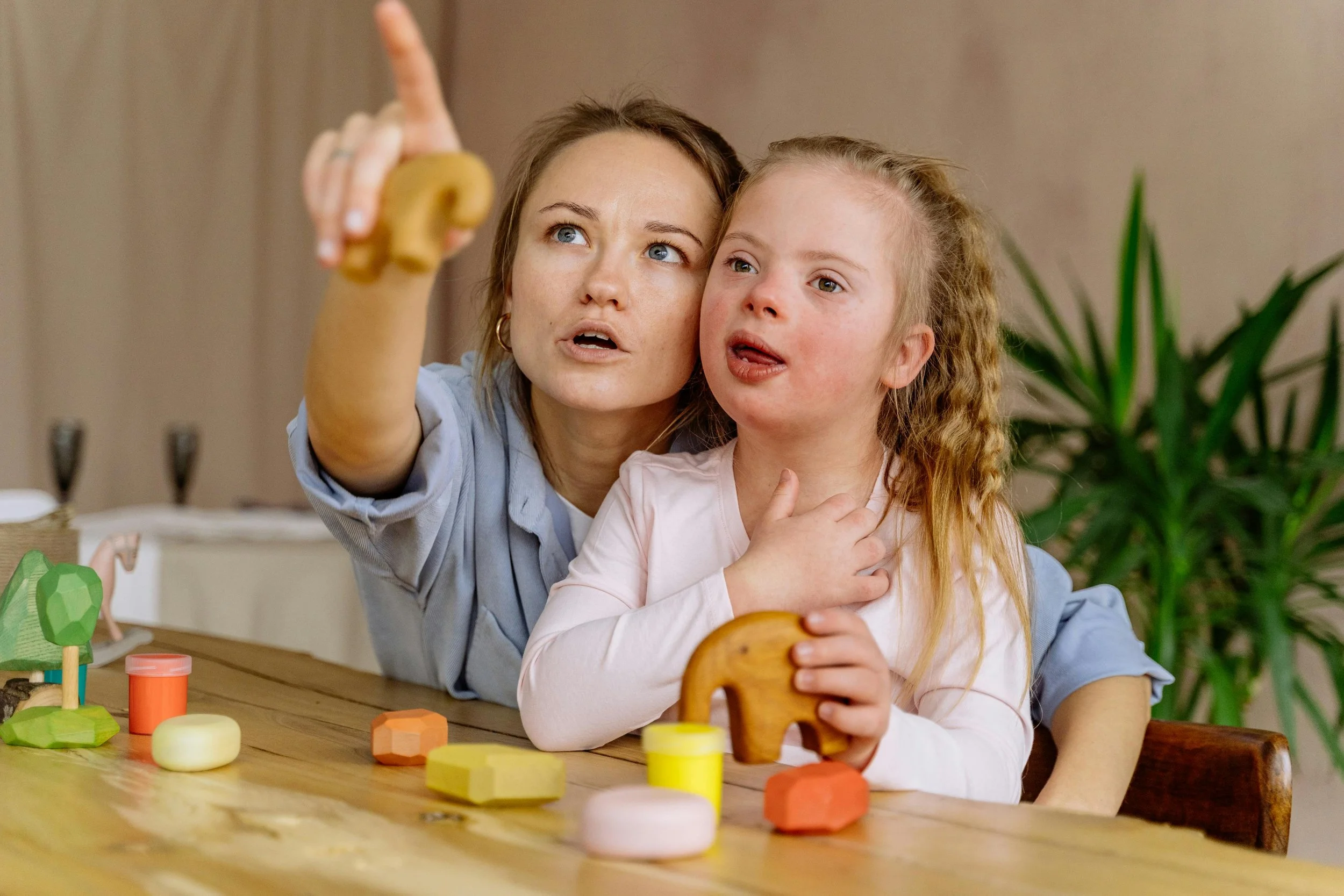 A woman and young girl sitting at a table with colorful wooden toys, the woman is pointing upwards as they look and talk together.