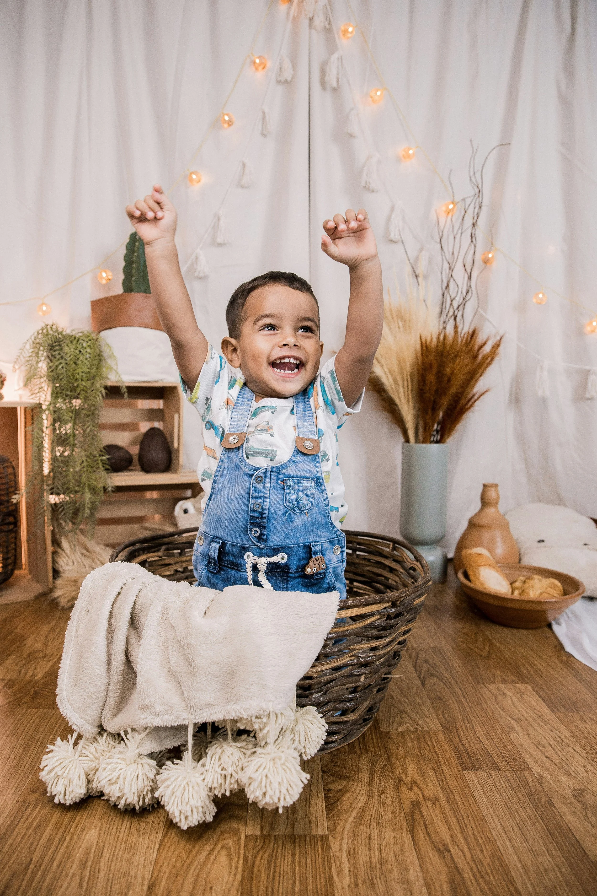 A happy child standing in a basket with arms raised, inside a room decorated with string lights, plants, and neutral-colored decor.