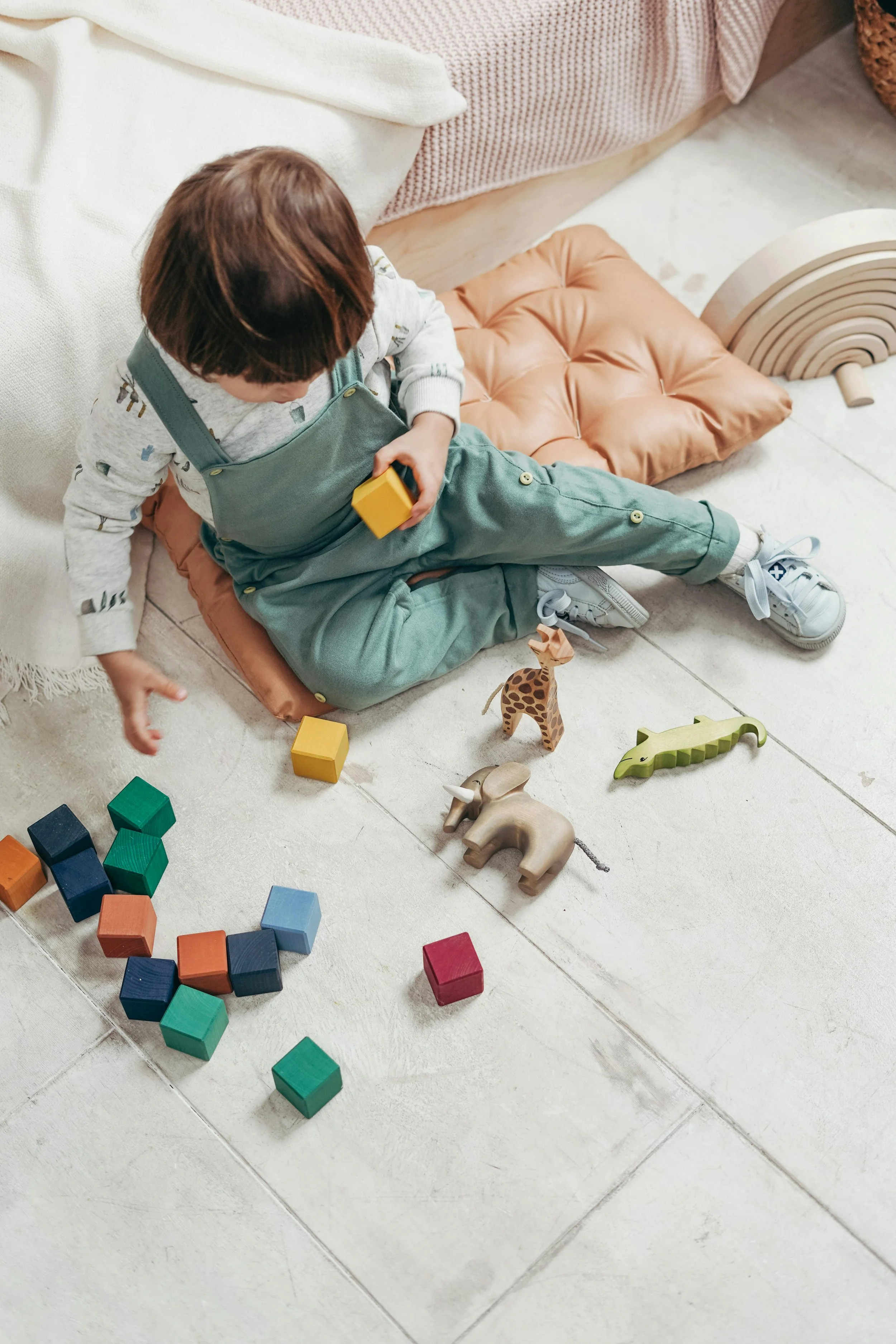 A young child sitting on a floor surrounded by colorful wooden blocks and toy animals, playing quietly indoors.
