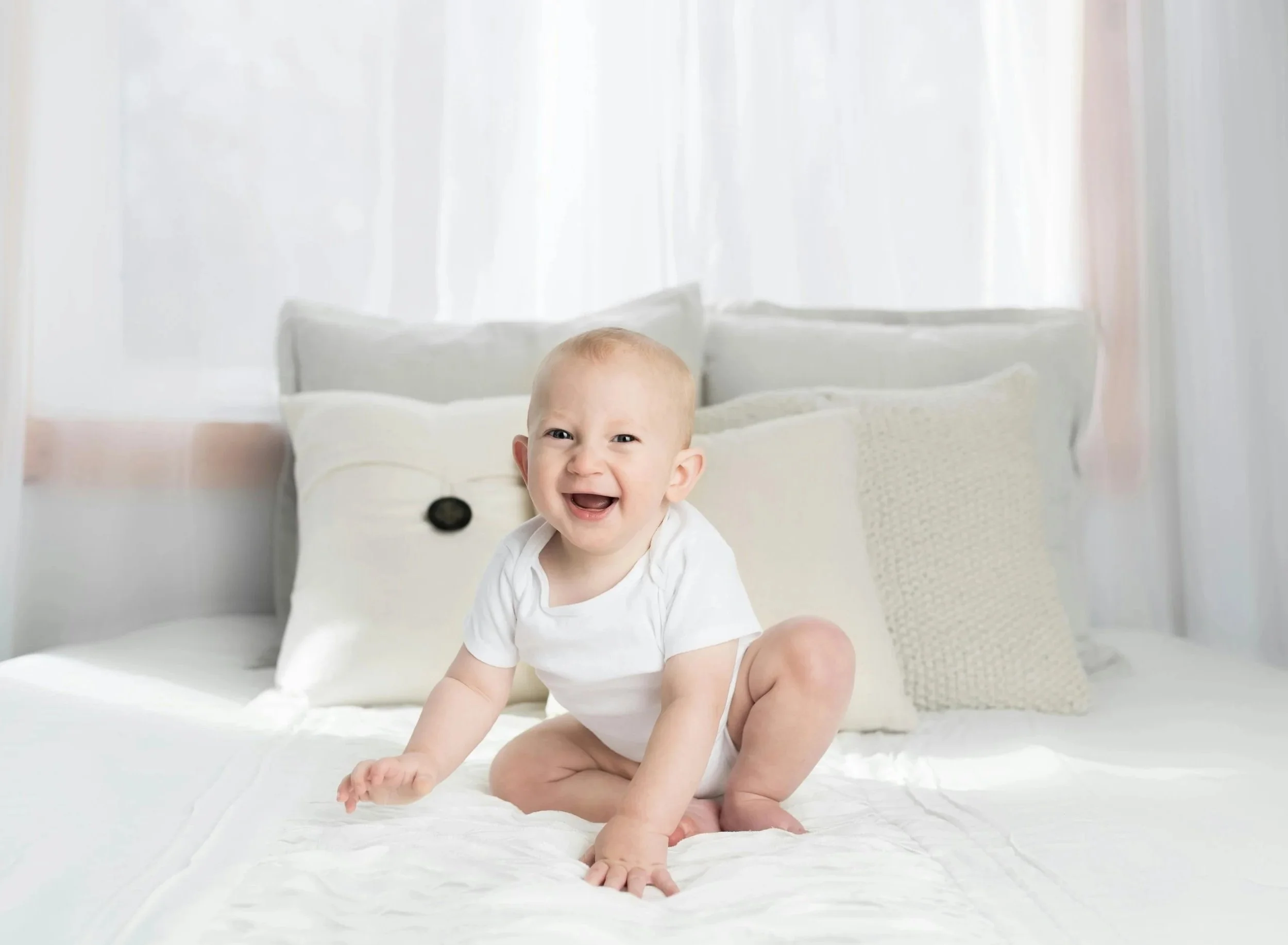 A smiling baby with light hair sitting on a bed with white sheets and pillows in a bright room.