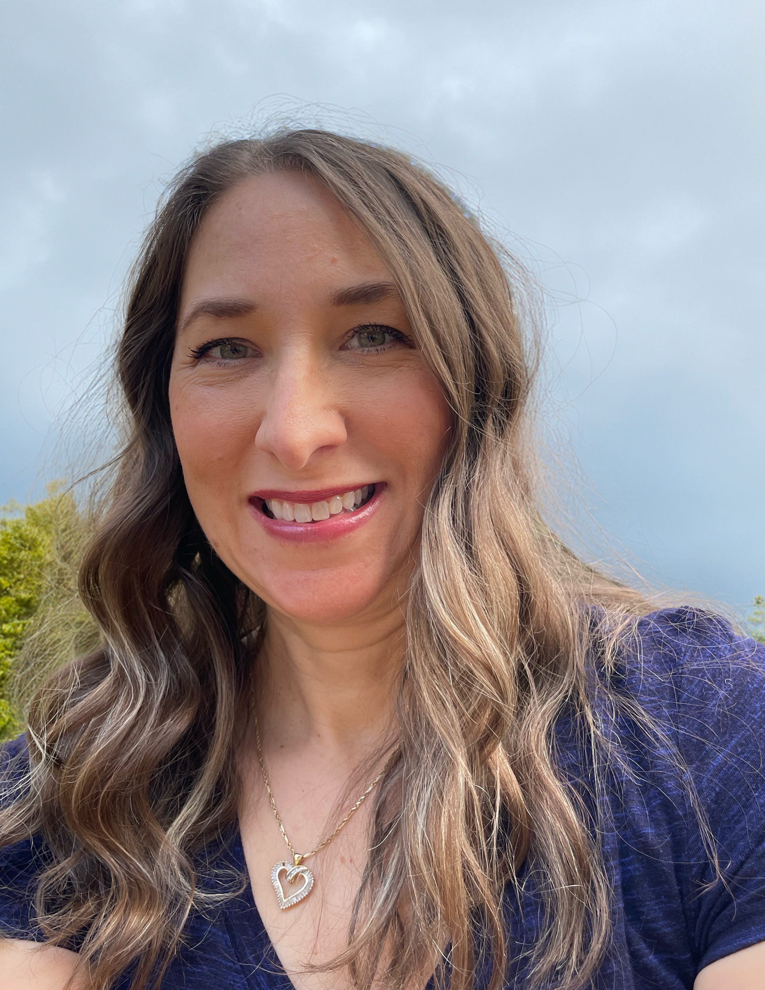 A woman with long wavy brown hair smiling outdoors with green trees and a cloudy sky in the background.
