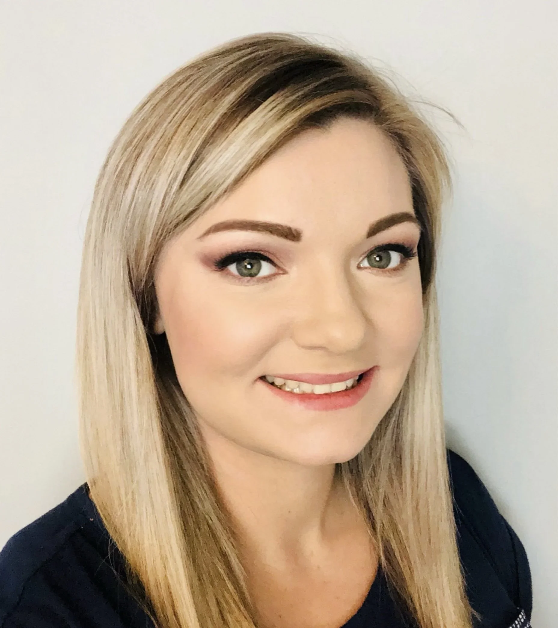 Close-up photo of a smiling woman with long blonde hair, wearing makeup, and a dark top.