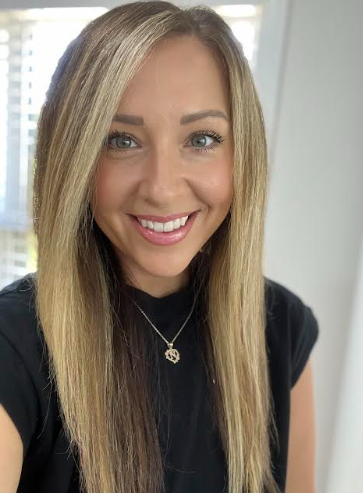 Portrait of a smiling woman with long blonde hair, blue eyes, wearing a black top and gold necklace, standing indoors near a window with city buildings in the background.