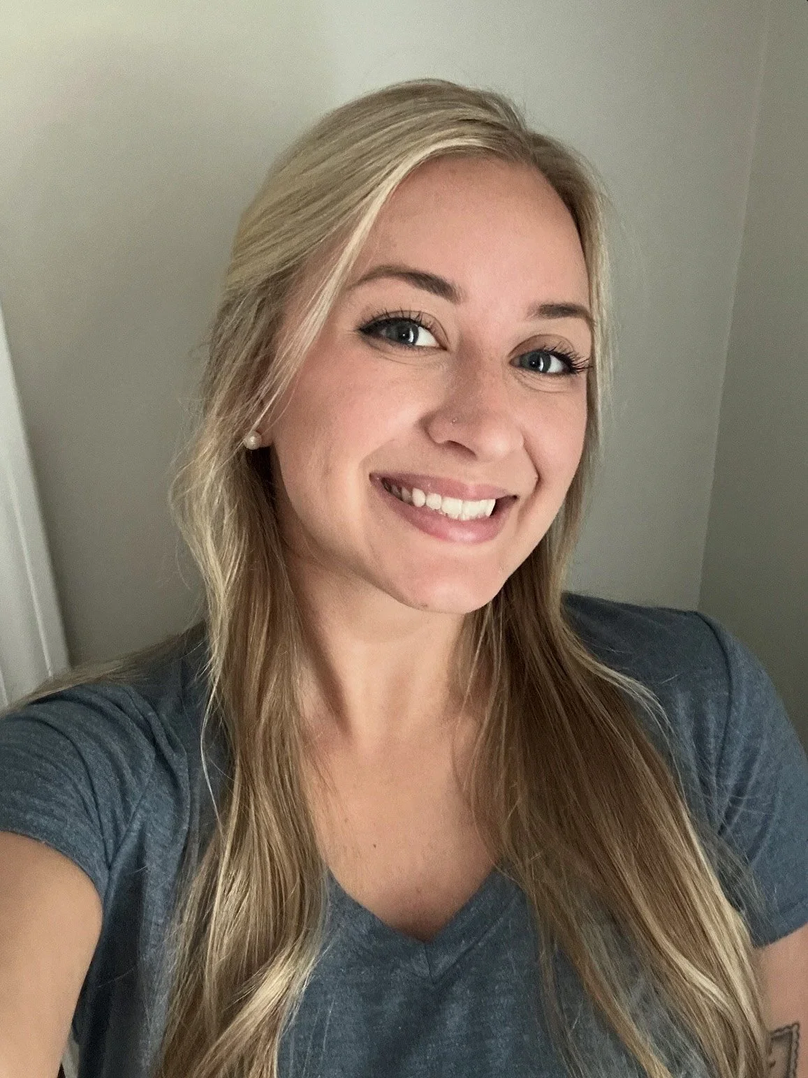 A smiling woman with long blonde hair, blue eyes, and pearl earrings taking a selfie indoors, wearing a dark gray t-shirt.