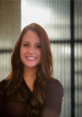 A woman with long brown hair smiling, standing indoors near a large window with vertical blinds.