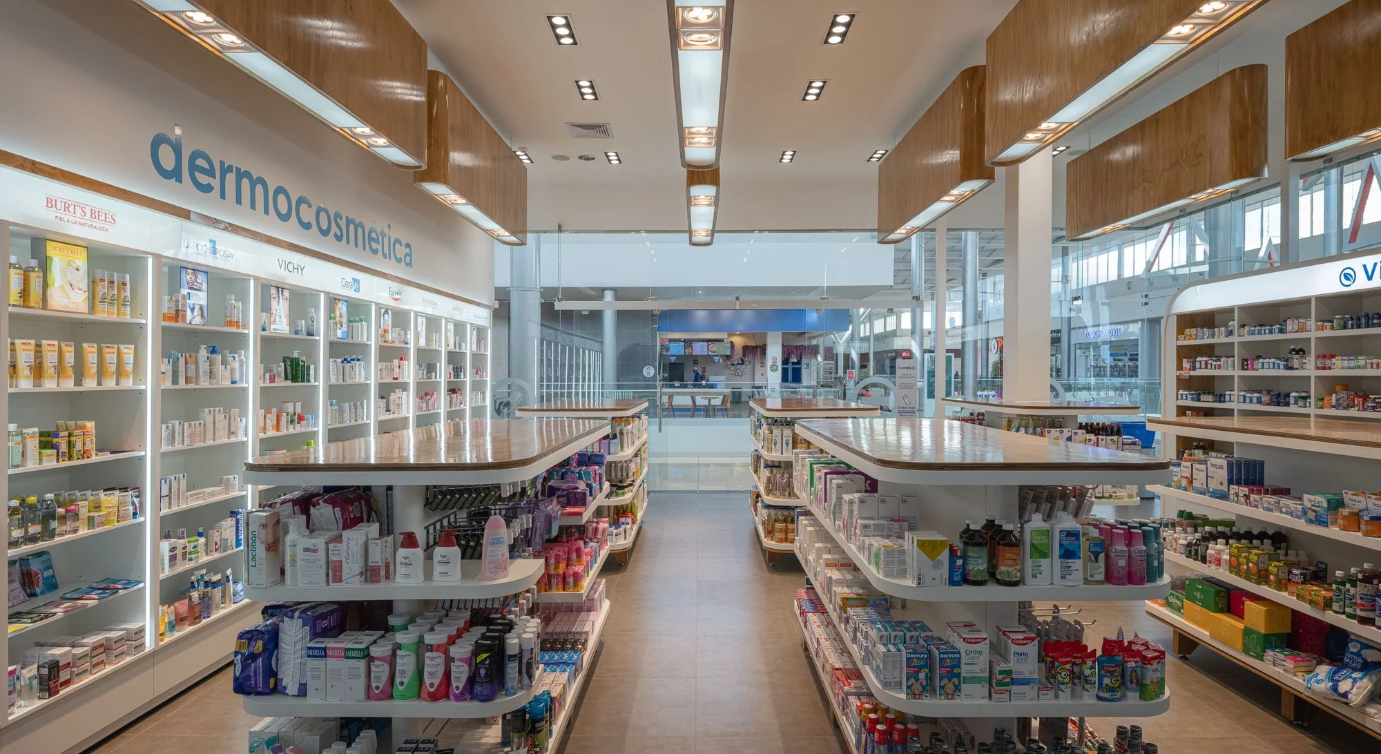 Interior de una farmacia con estantes llenos de productos para cuidado de la piel, medicamentos y otros artículos de farmacia, con una área de caja al fondo y un ambiente moderno con mucha luz natural.