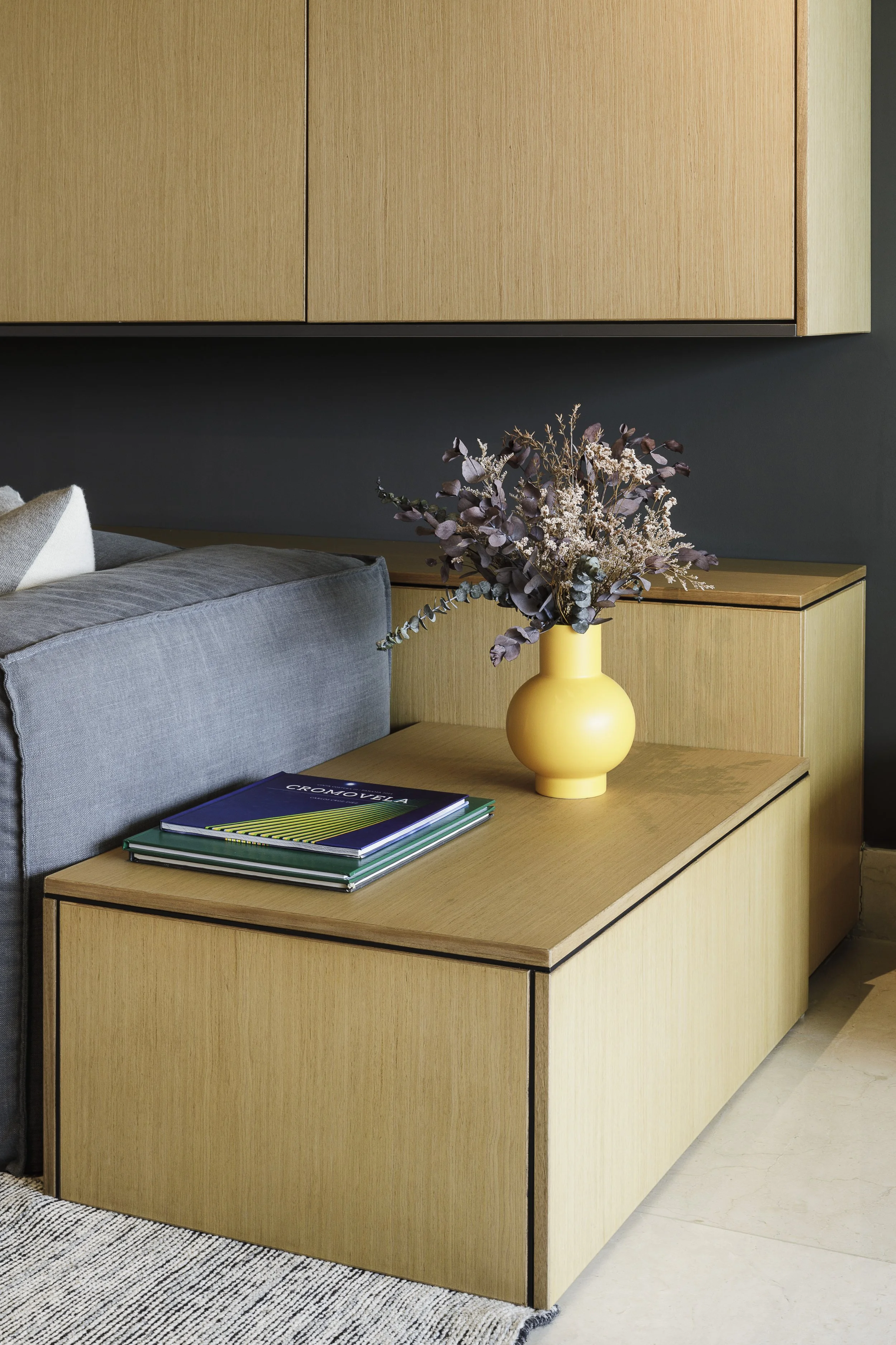 Mesa de madera con una pila de libros, una maceta amarilla con flores secas y una esquina de un sofá gris. Fondo con armario de madera y pared gris oscuro.