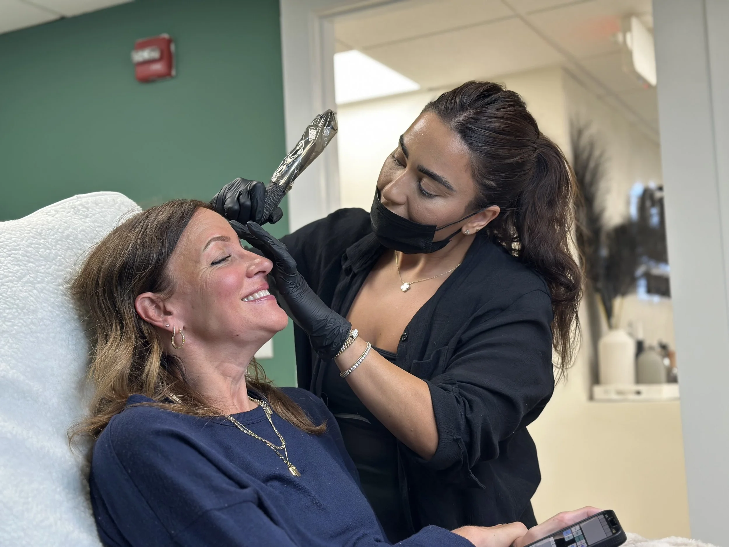 A woman lying in a chair receiving a cosmetic treatment on her forehead by a professional wearing a black mask and gloves in a modern clinic.