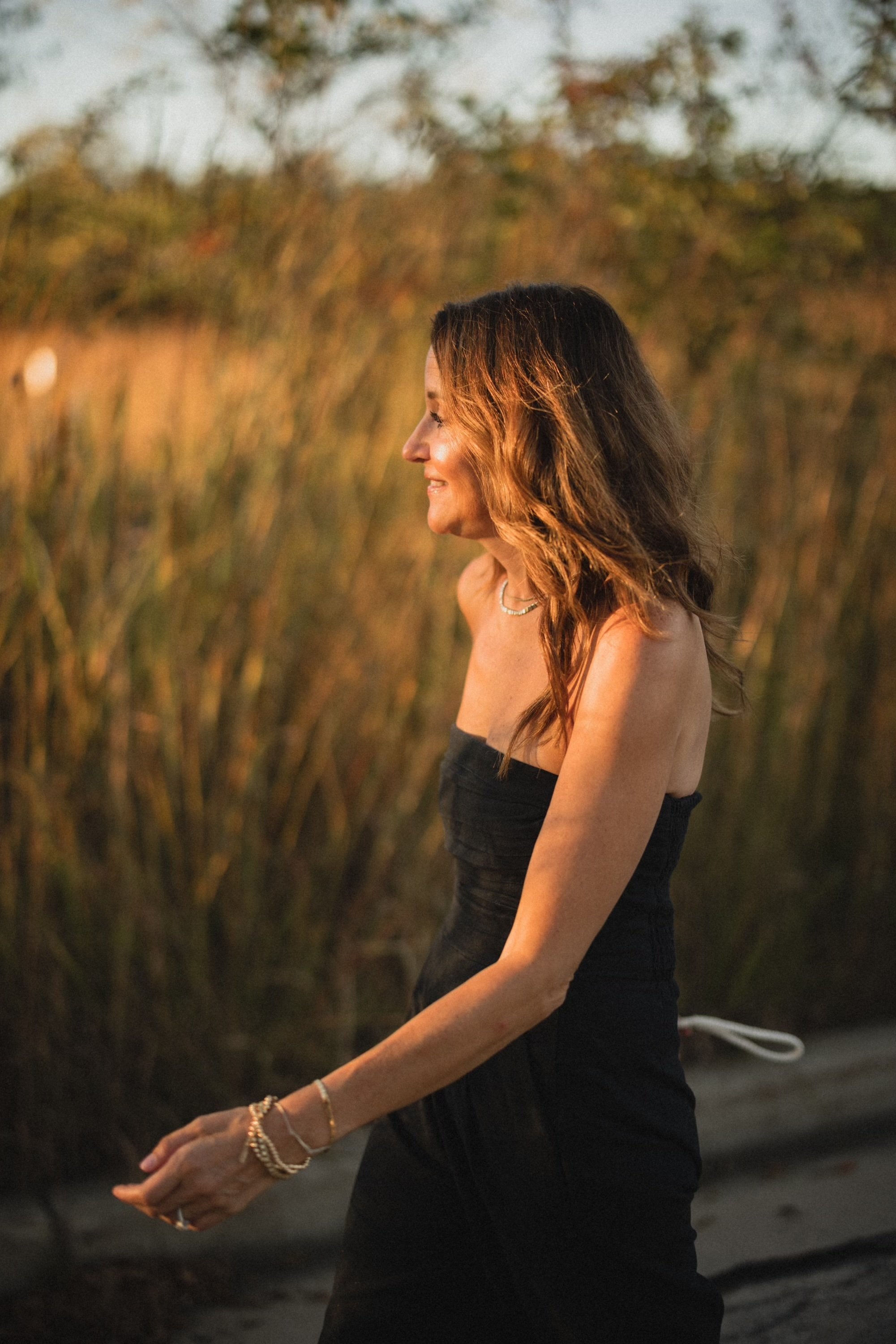 A woman with brown hair wearing a black strapless dress stands outdoors at sunset, smiling and looking to the side, with trees and bushes in the background.