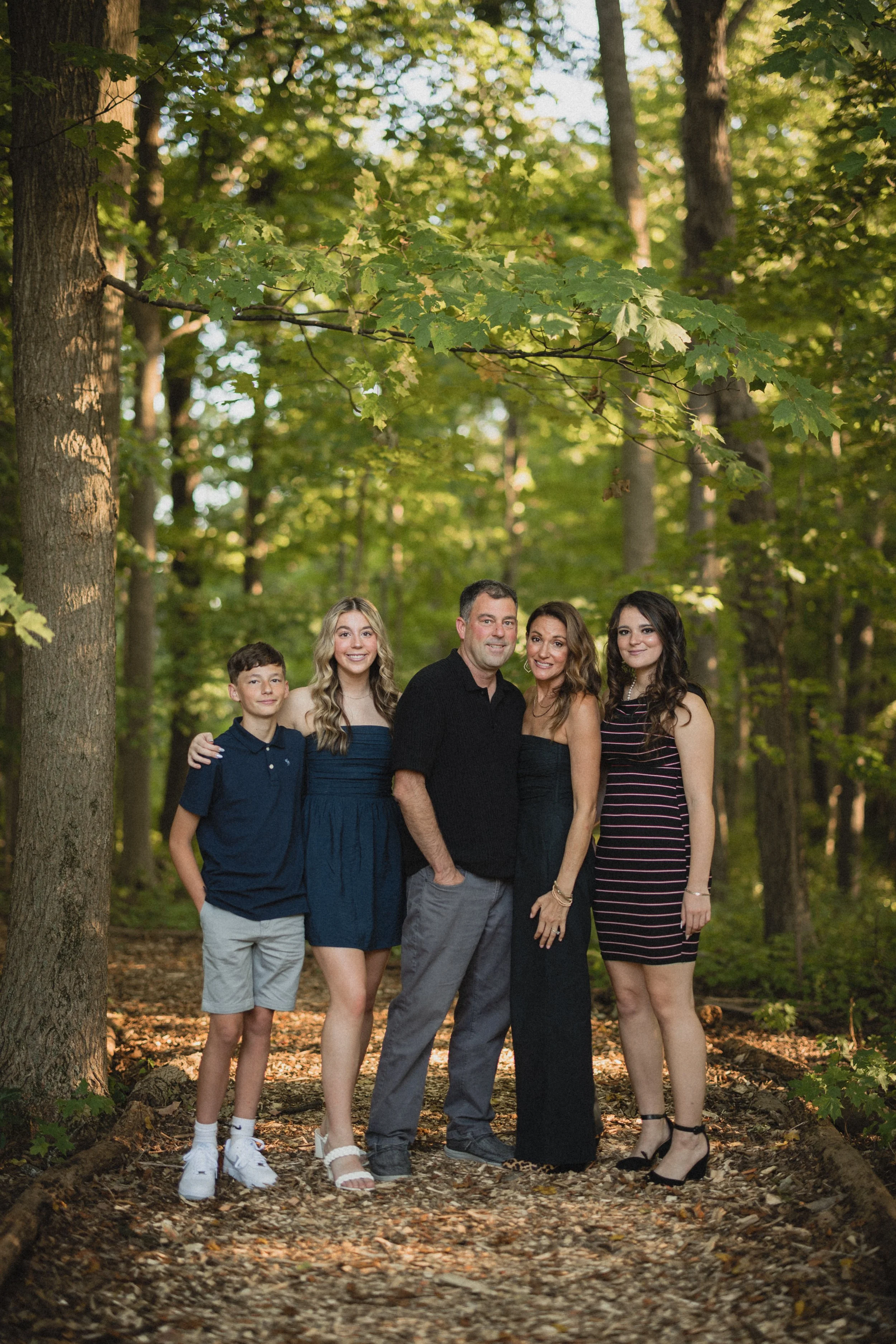 A family of five standing on a wooded trail with green foliage in the background, smiling at the camera.
