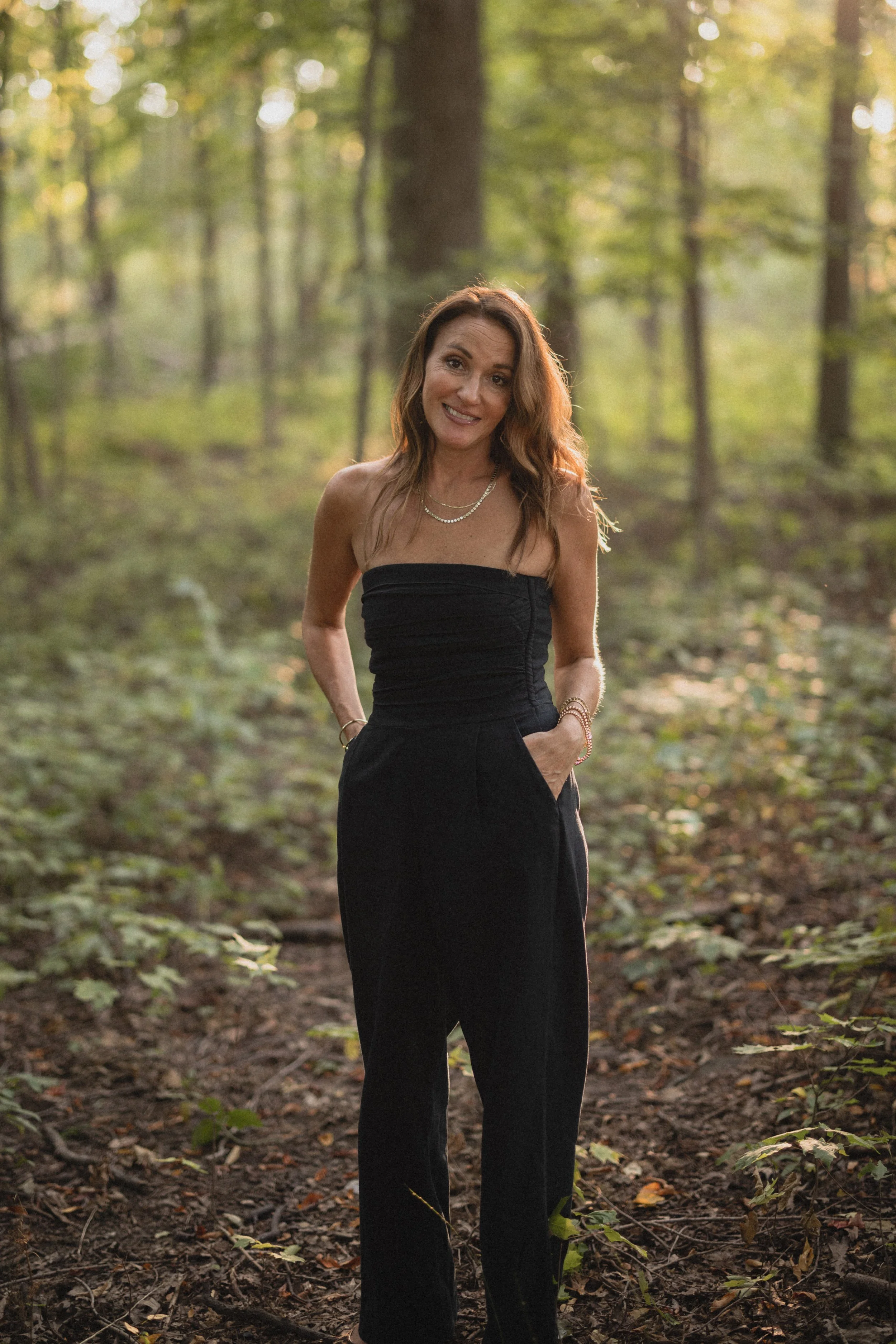 A woman in a black strapless jumpsuit standing with her hands in her pockets outdoors in a wooded area during daylight.