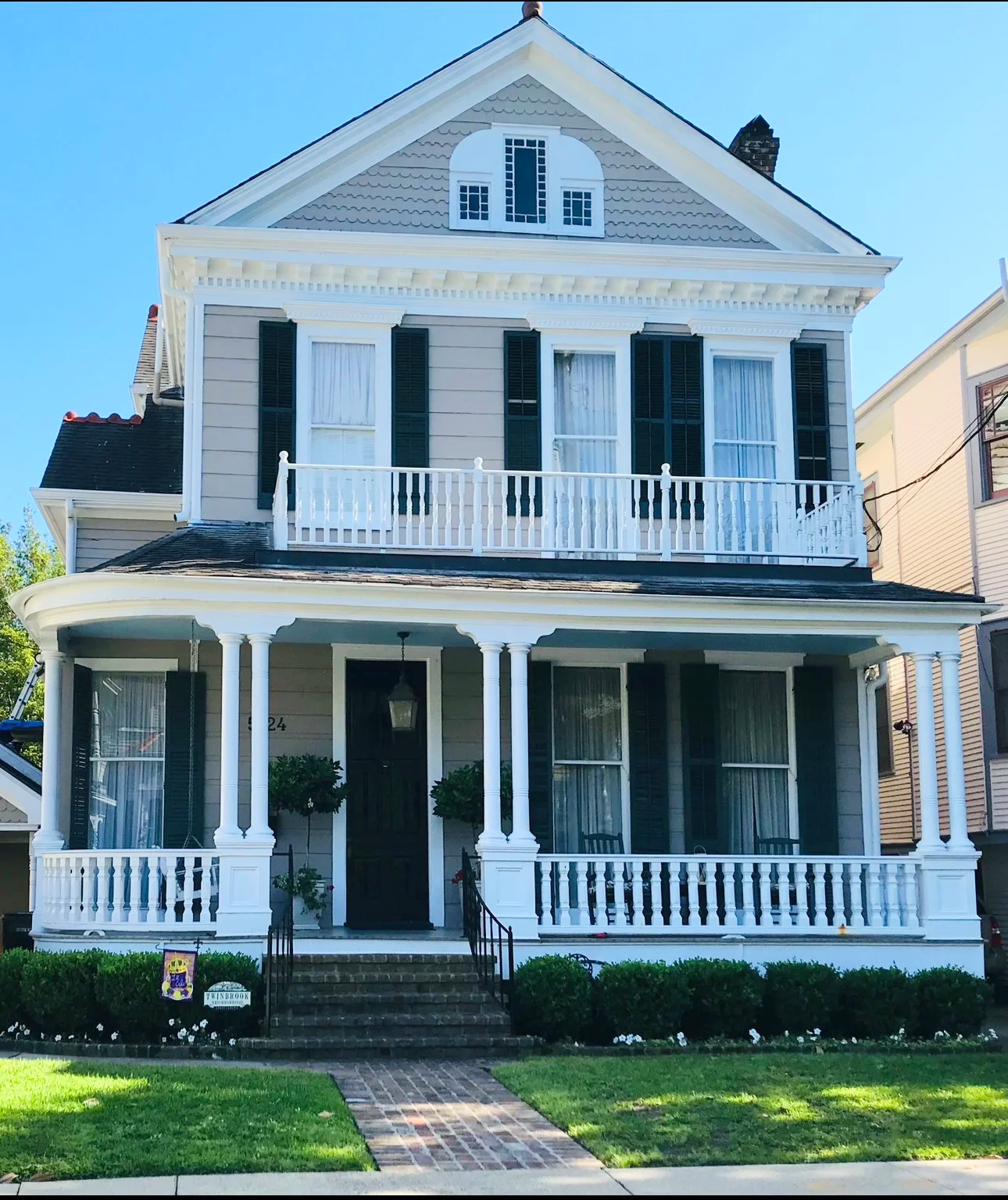 A three-story Victorian house with gray siding, black shutters, and a white porch with columns, and a second-floor balcony, set against a clear blue sky.