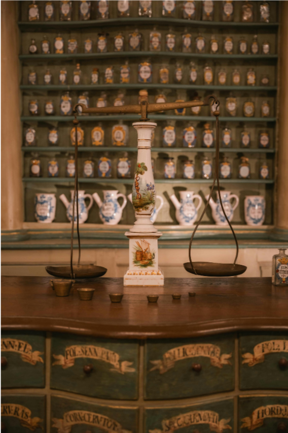 Antique balance scale on wooden counter with shelves of labeled jars and pitchers behind.