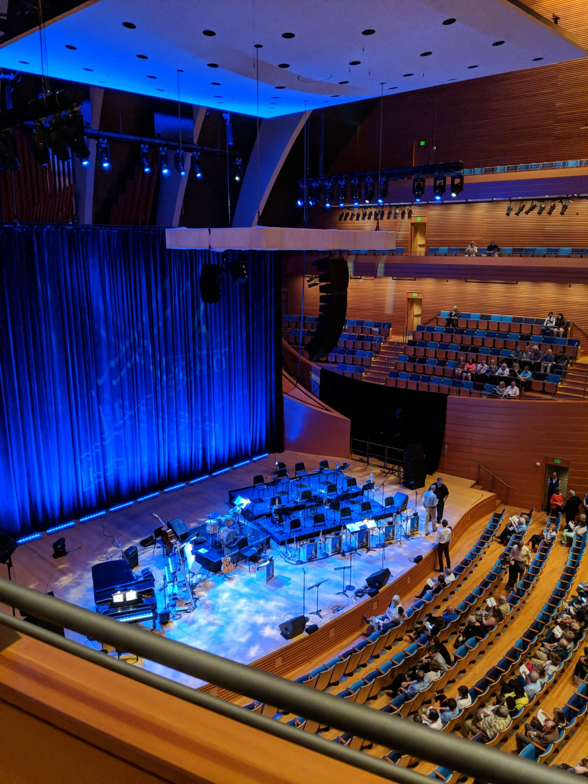 Interior view of a concert hall with a stage set up with musical instruments, blue lighting, and an audience seated in the auditorium, taken from an upper balcony.