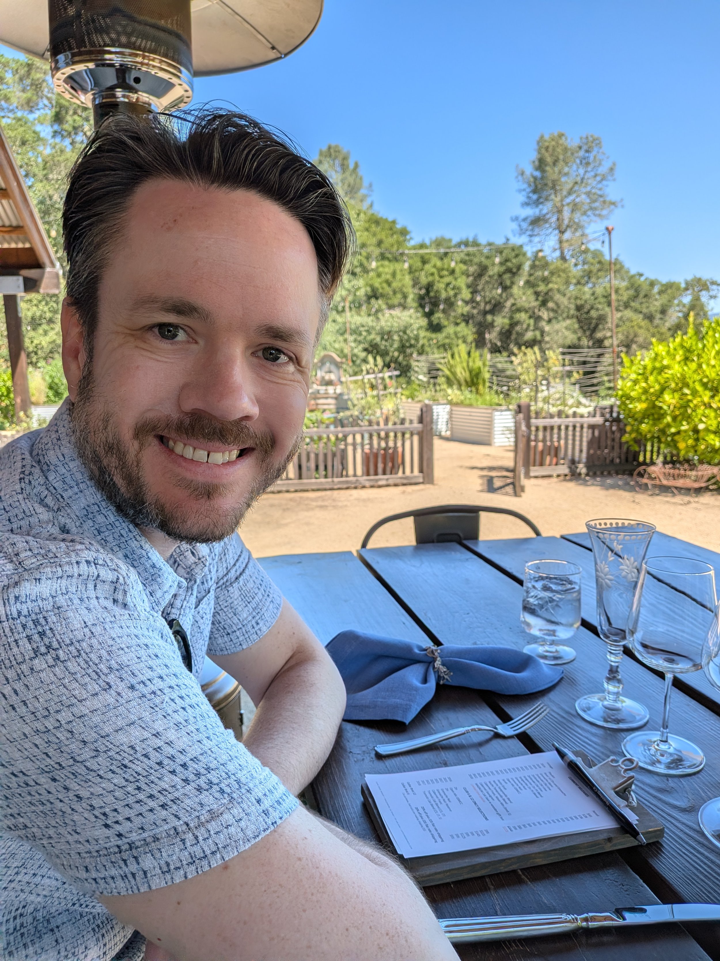 A man smiling at a table with wine glasses and trees in the background at Capo Creek Winery in Healdsburg, California