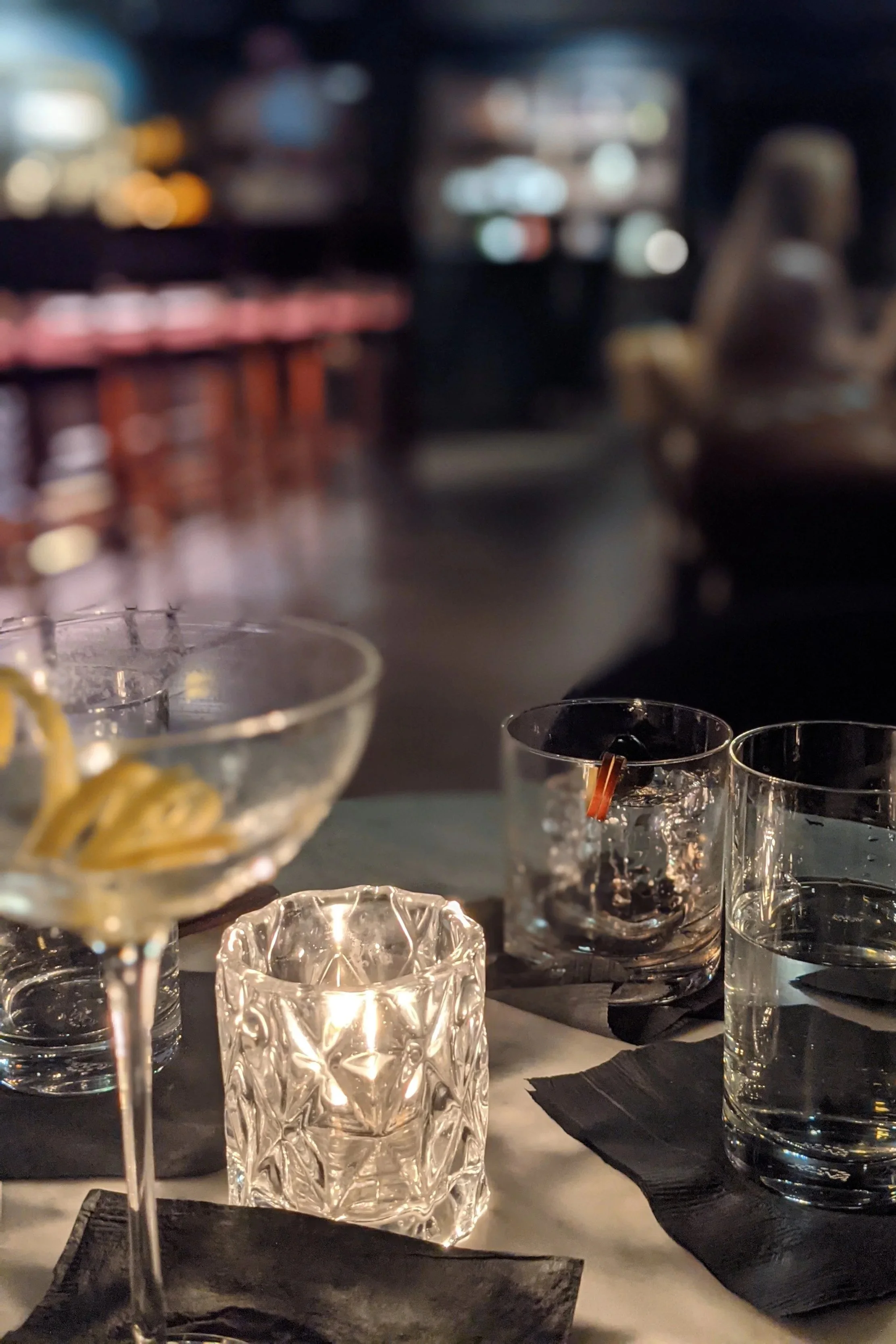 A dimly lit restaurant table with several glasses of water and a glass of champagne, black napkins, and a dark tablecloth, with blurred background of bar or restaurant interior with hanging lights.