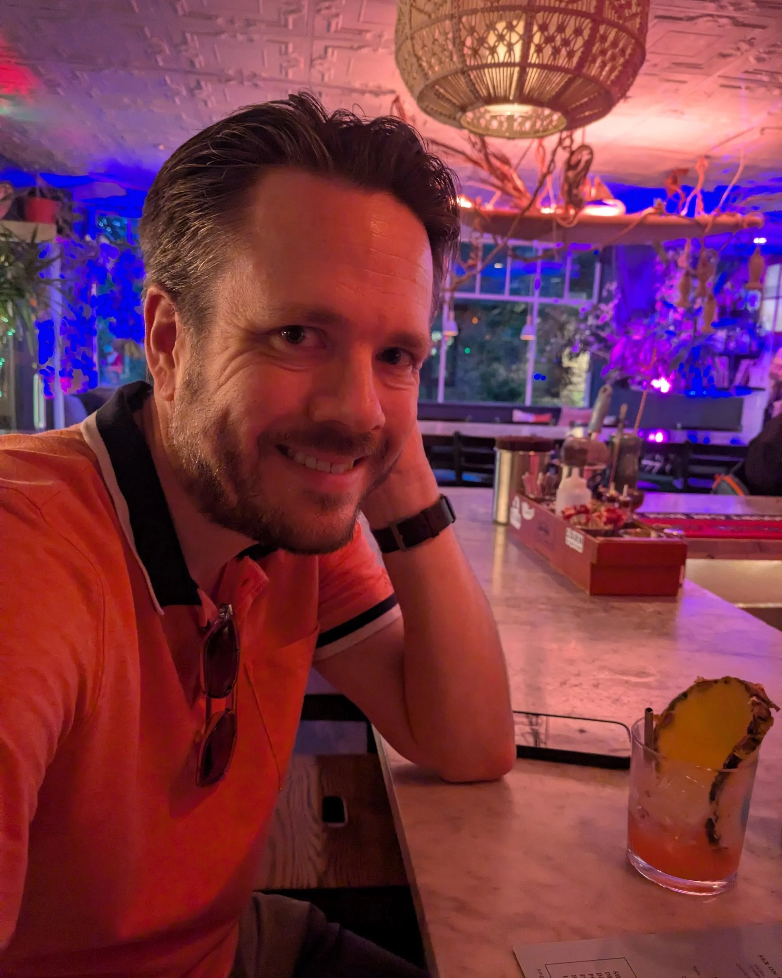 Man in a colorfully lit bar with a drink that has a slice of pineapple in the glass.