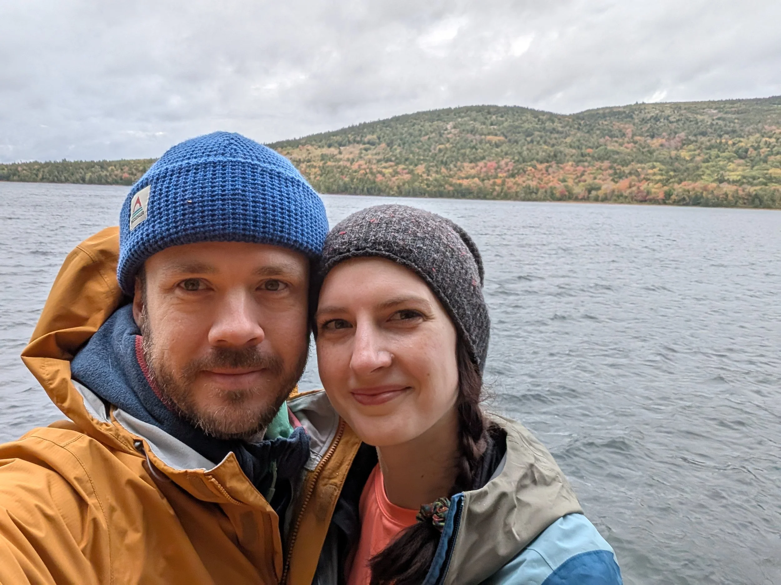 A man and a woman in rain jackets and beanies standing in front of a lake with trees of various colors in the background.