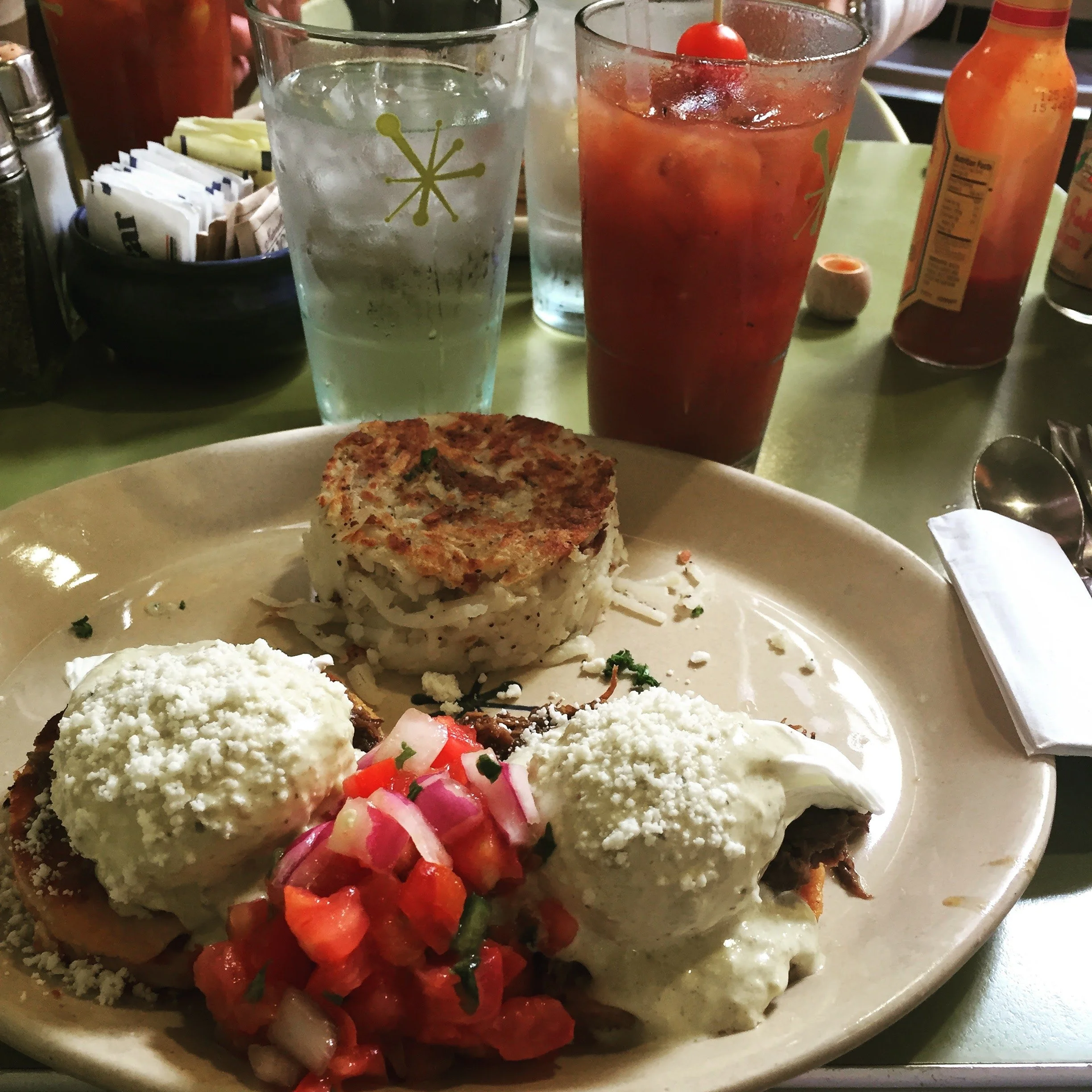 Plate of hash browns and eggs benedict with a glass of water and bloody mary in the background.