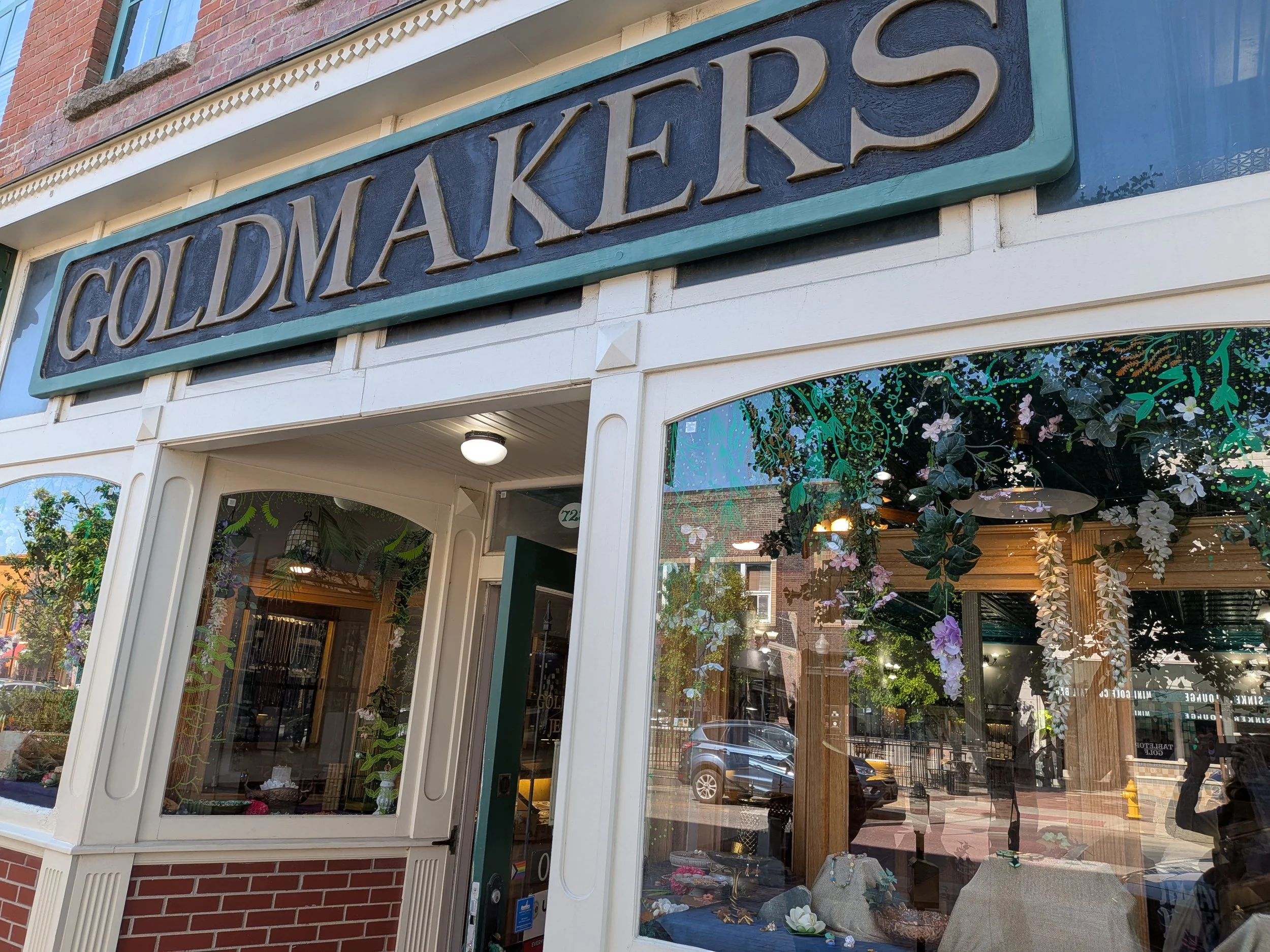 Storefront with a sign that says 'GOLDMAKERS' and large display windows decorated with plants and flowers.