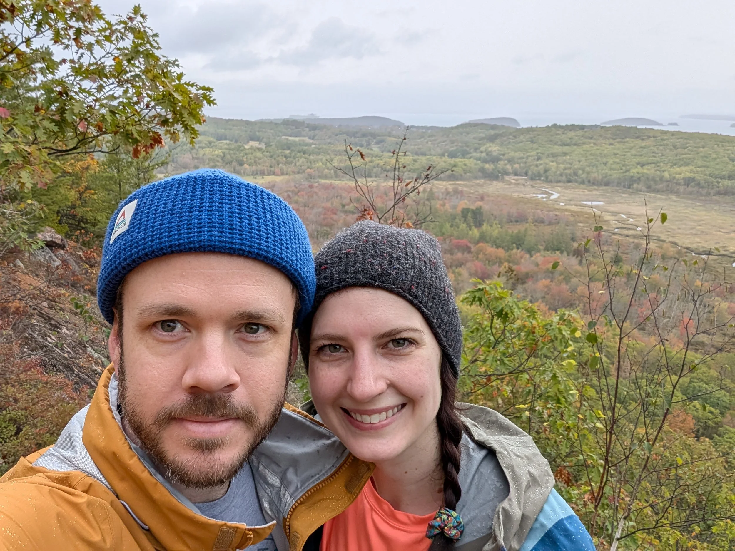 A man and a woman wearing beanies with a background of trees, marshland and ocean.