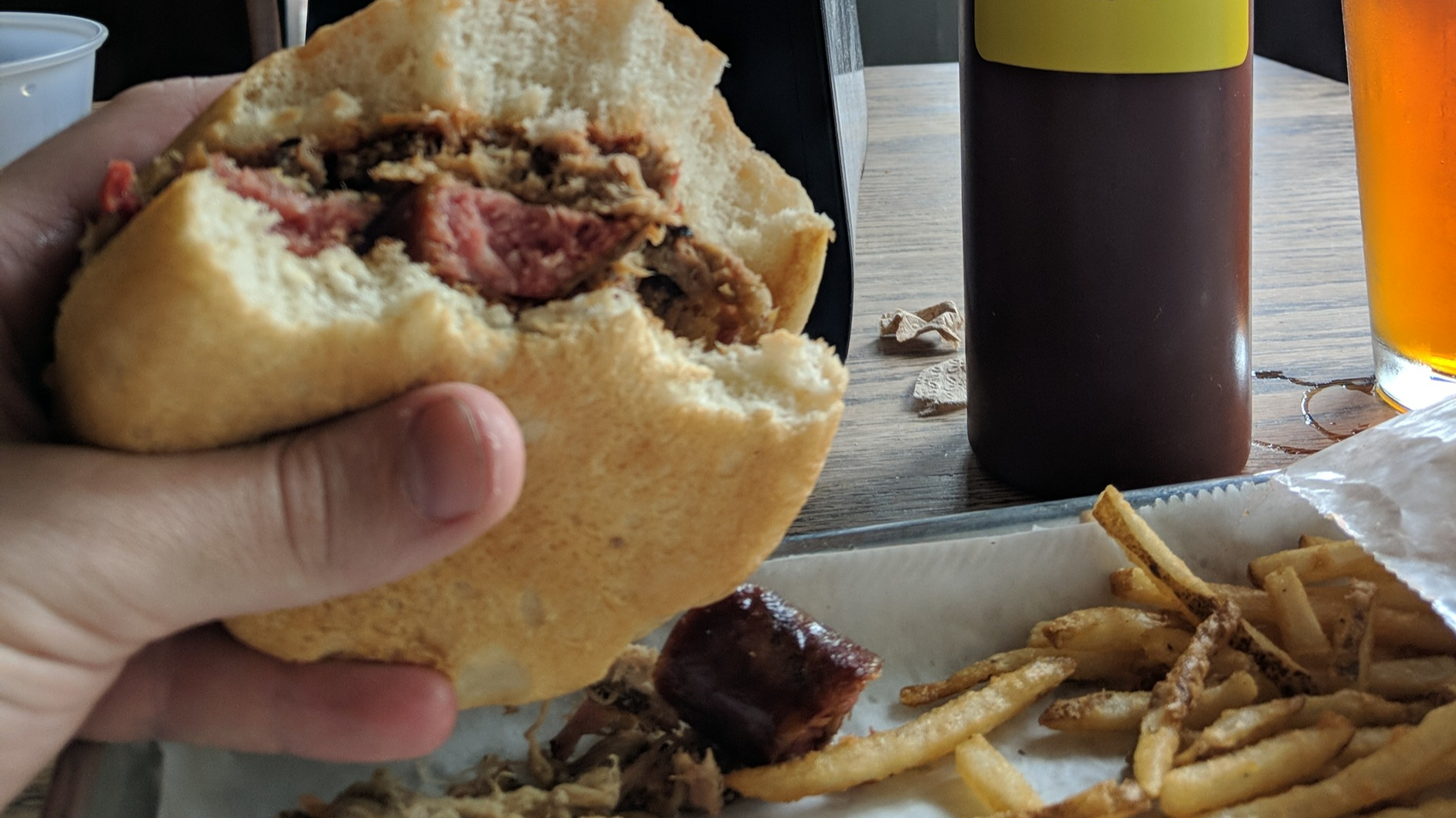 A hand holding a barbeque sandwich with a few bites taken over a tray with french fries.