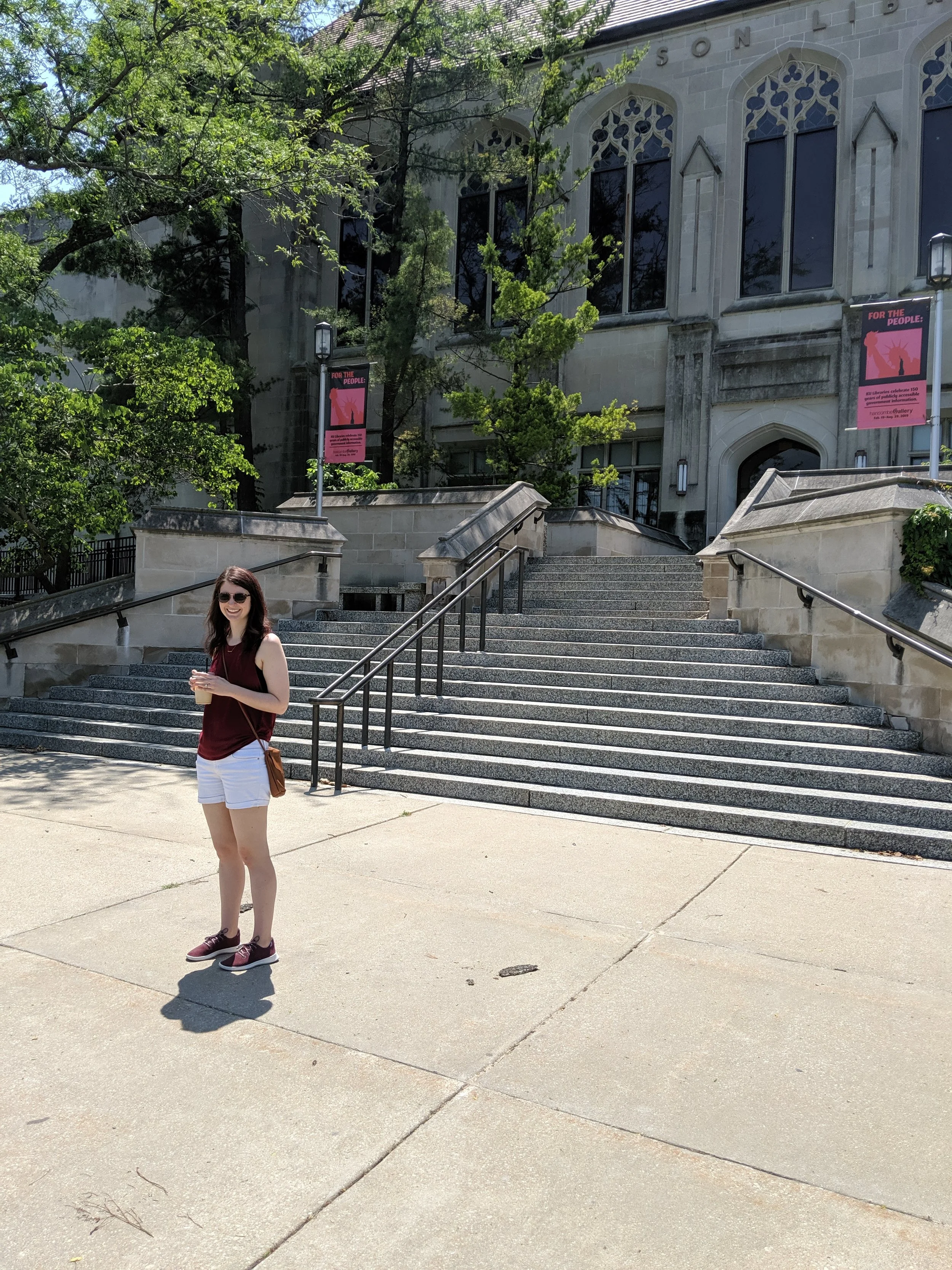 A young woman with sunglasses, a maroon sleeveless top, white shorts, and maroon sneakers, standing on a concrete sidewalk in front of a staircase leading to a historic stone building, with trees and banners around.