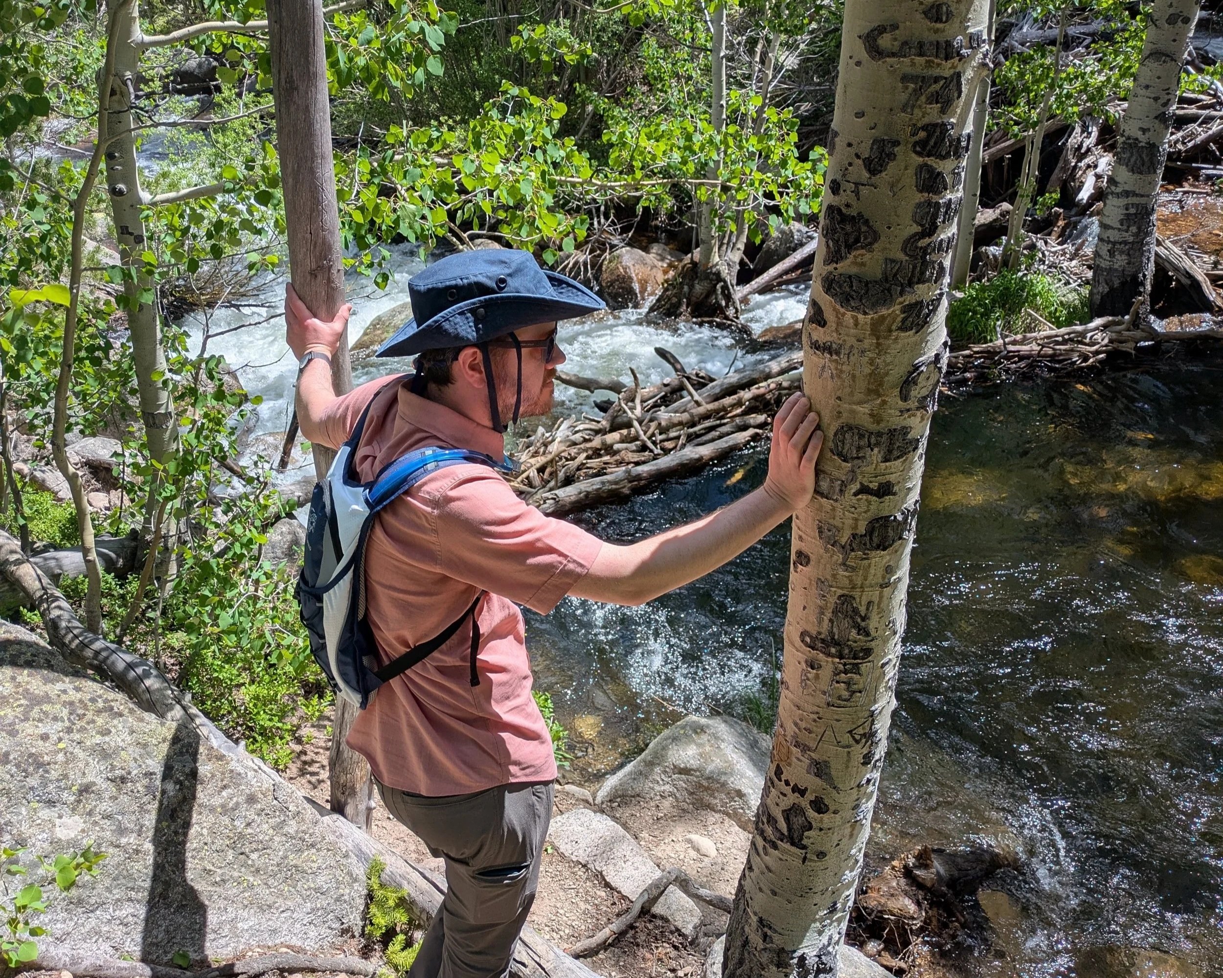 Man in a hiking outfit standing next to a river looking out  with his hands on two trees.