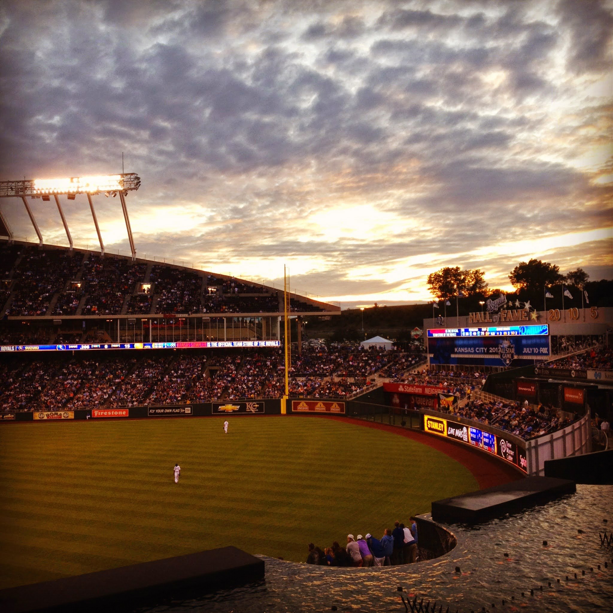 A baseball stadium filled with spectators during sunset, with the field visible and some players on it. The sky is partly cloudy with the setting sun casting a warm glow.