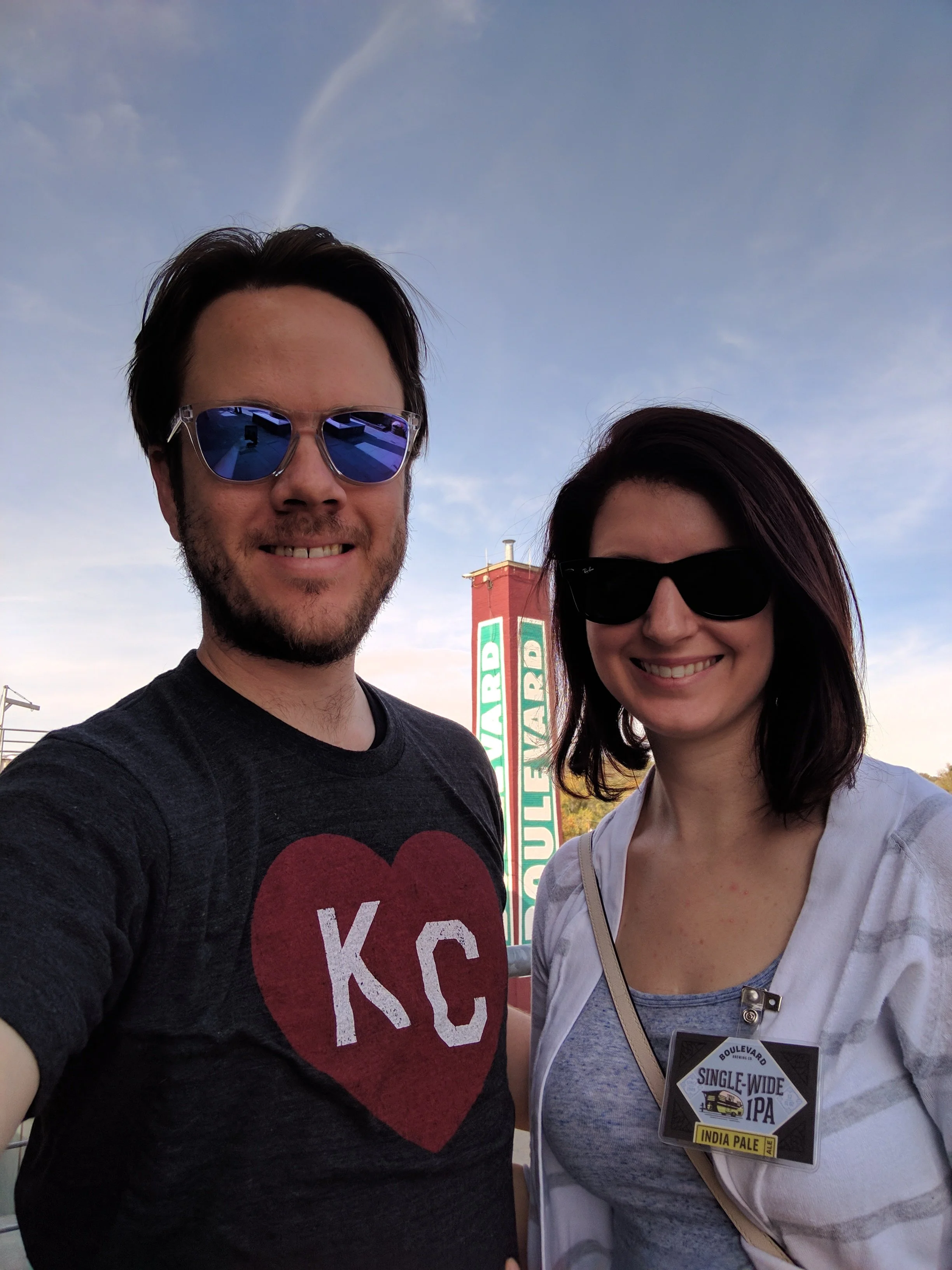 A man and woman wearing sunglasses taking a selfie outdoors during daytime, with a sign in the background advertising Boulevard Single-Wide IPA India Pale Ale.