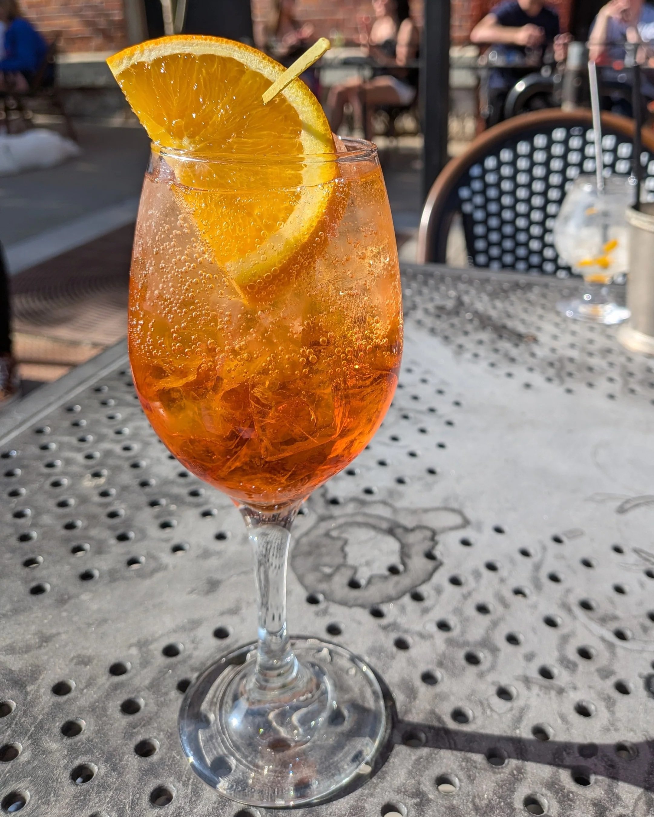 An aperol spritz in a wine glass with an orange slice on a metal table with chairs in people in the background.