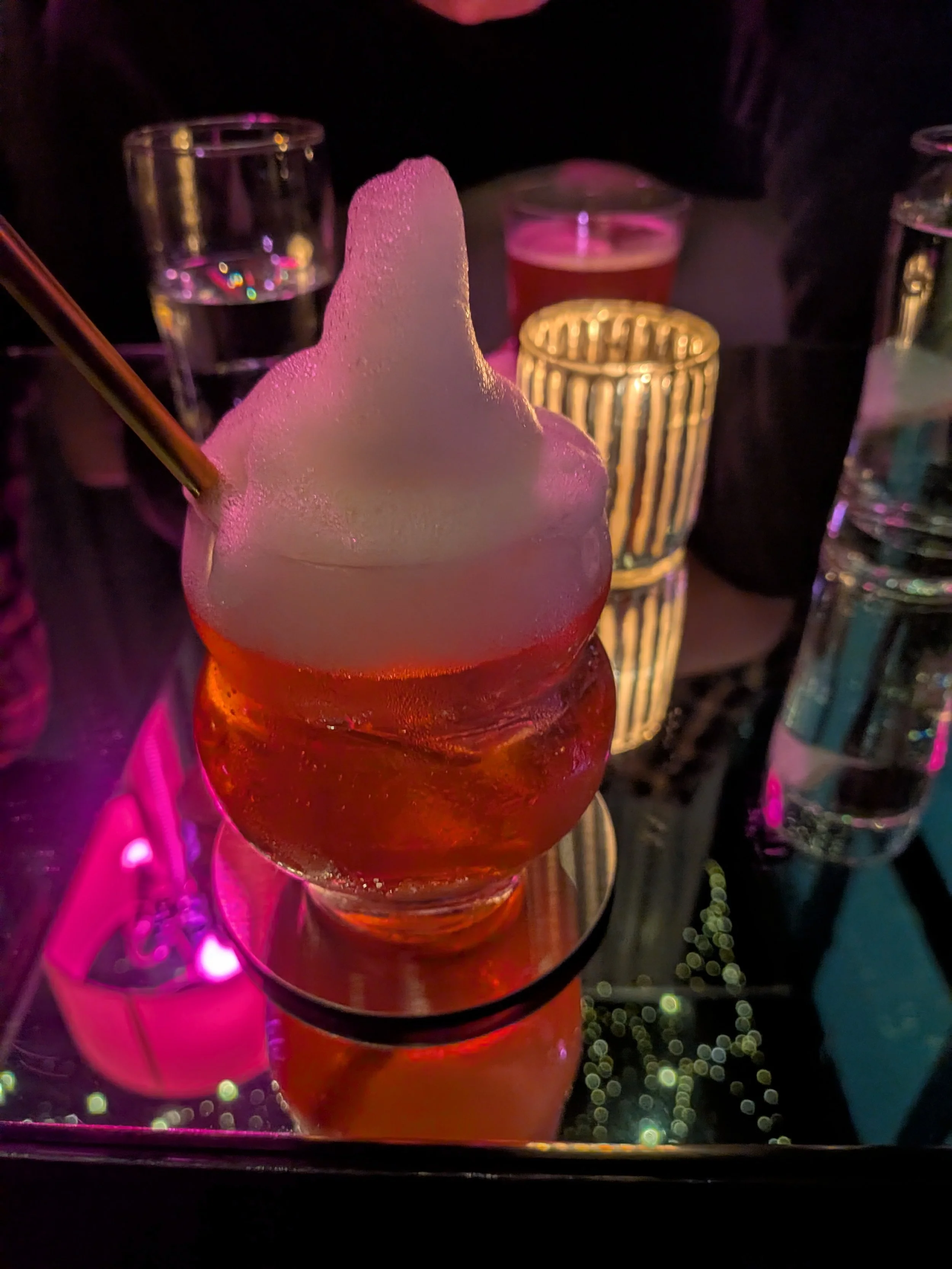 A glass with red-colored cocktail and large foam ice cream on top, with a metal straw. In the background, there are several glass cups and decorative lights.
