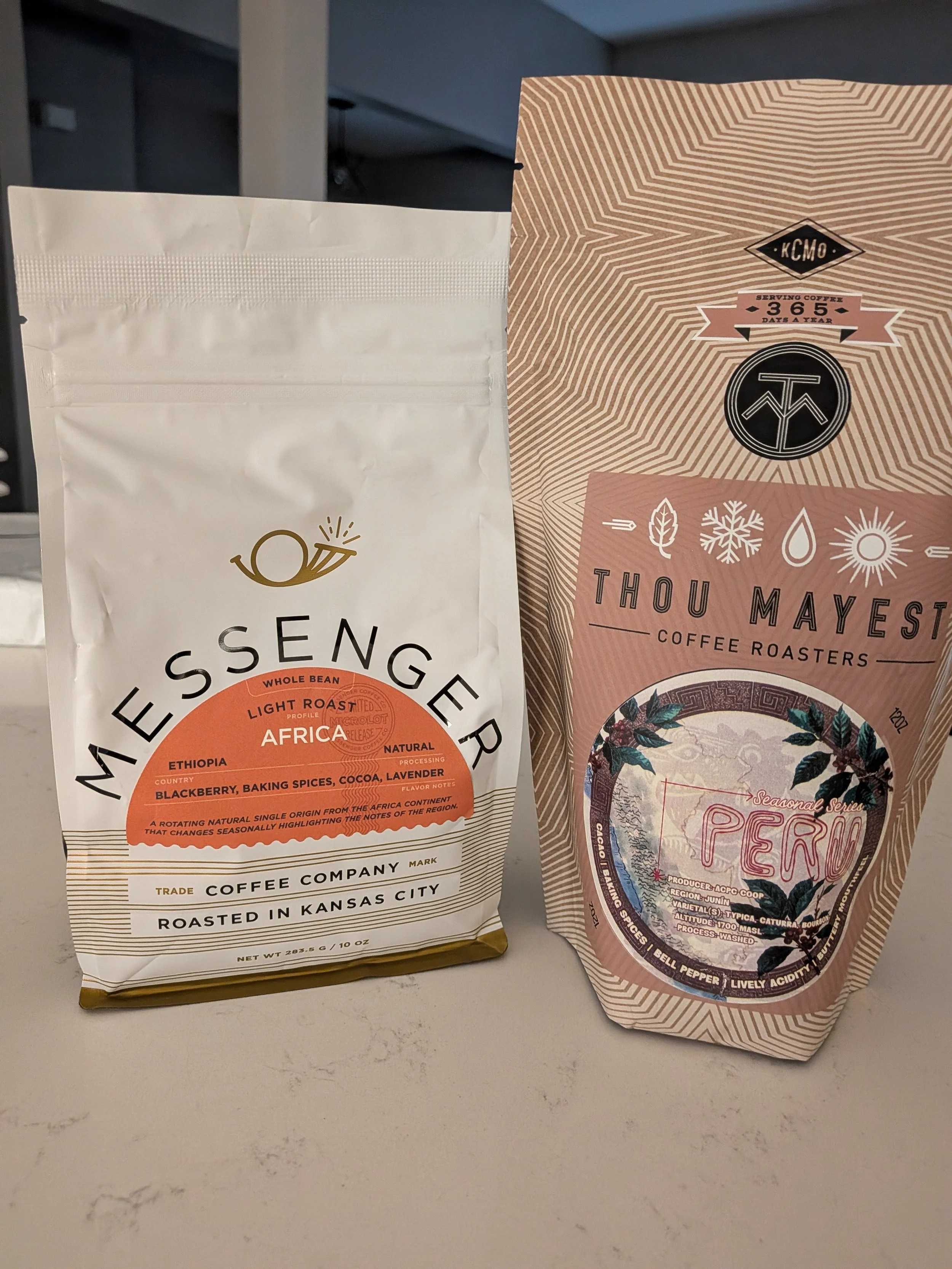 Two coffee bags on a white surface. The bag on the left is from May Seng in Africa, highlighting Ethiopia, Kenya, and other regions with notes on flavor profiles. The bag on the right is from Thou Mayest Coffee Roasters, featuring a colorful label with images of mountains and flora.