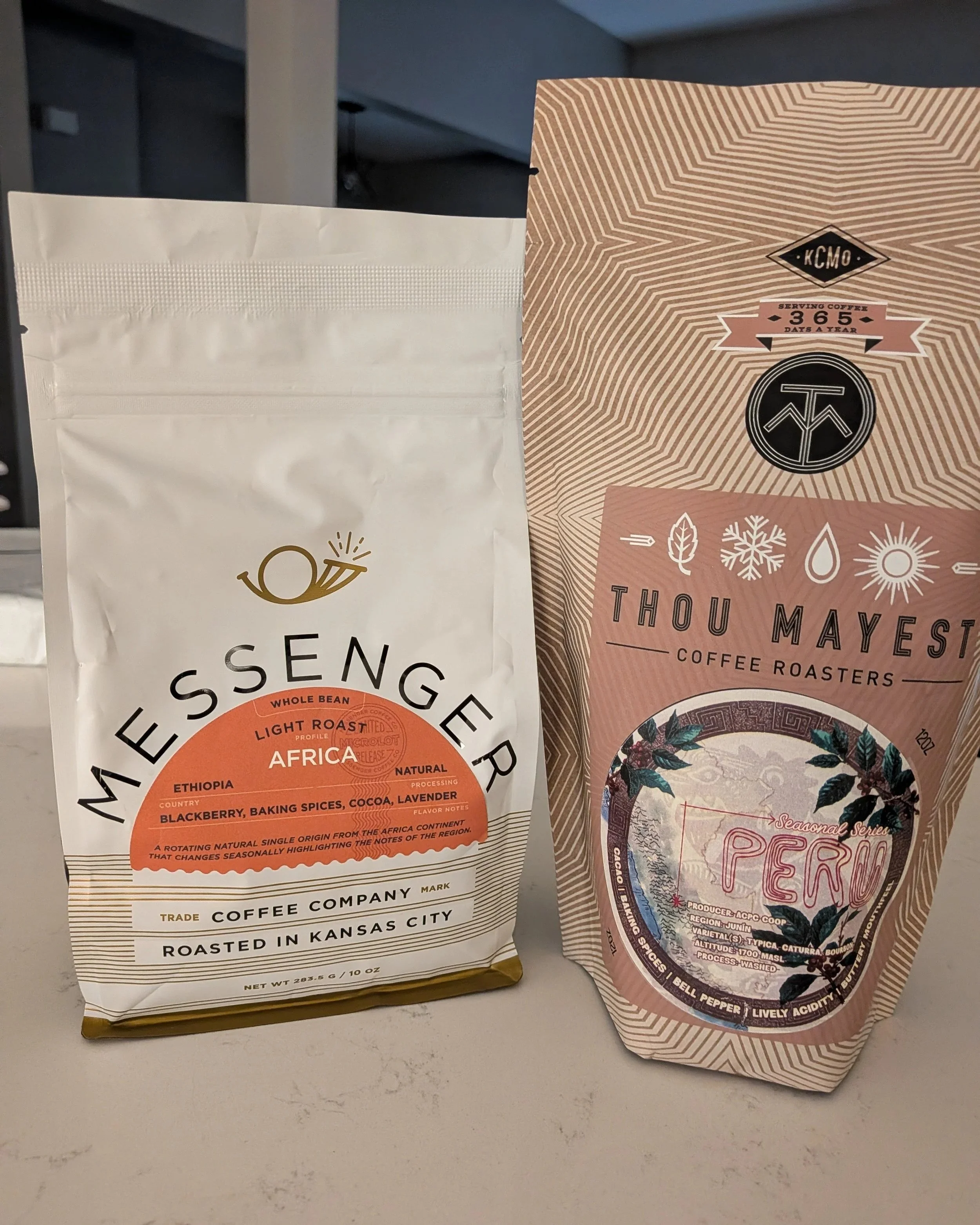Two bags of coffee beans from Messenger Coffee and Thou Mayest Coffee Roasters sitting on a white counter.