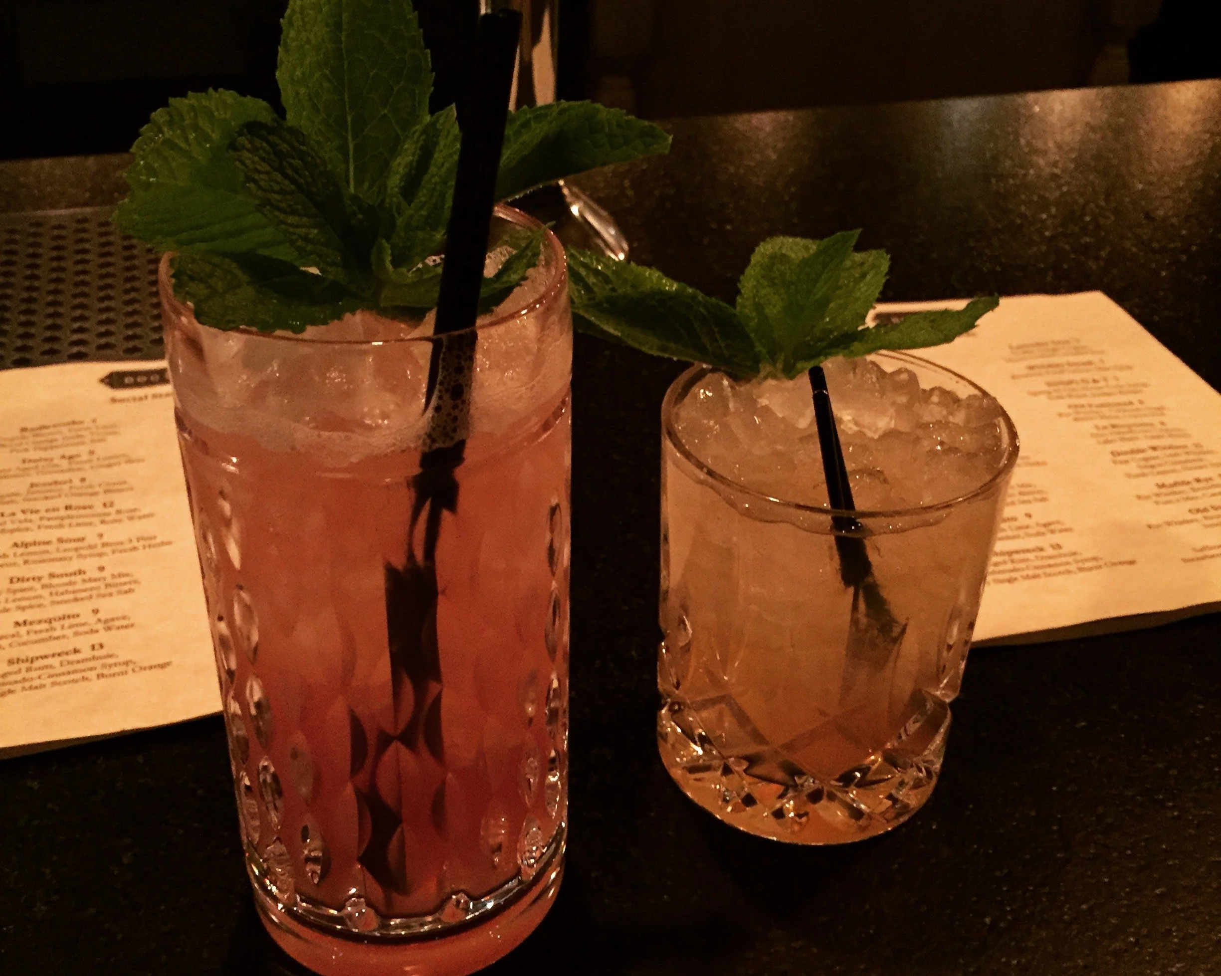 Two cocktails with strawas and mint garnishes on a table with menus on the table behind them.