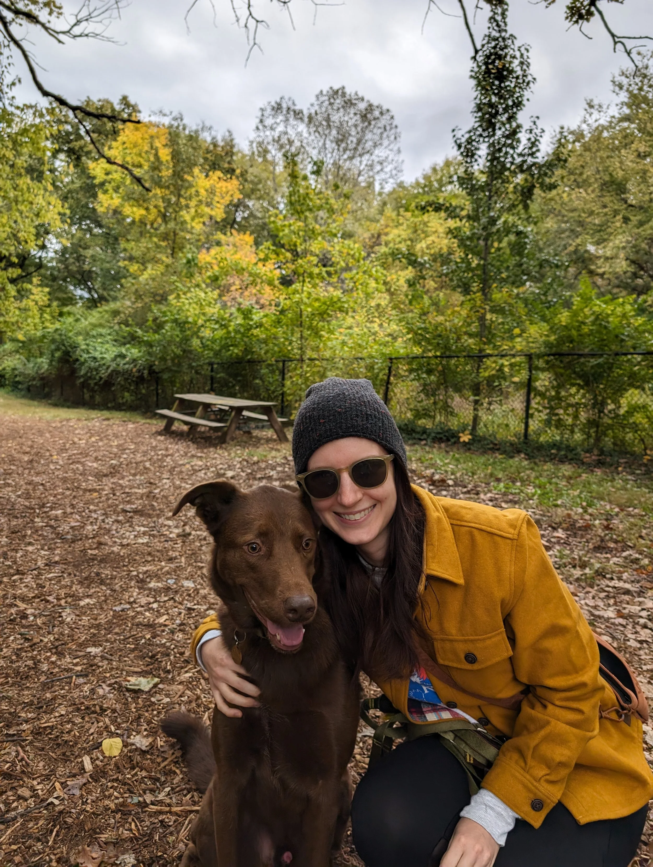 A woman wearing sunglasses and a gray knit hat is smiling while crouching next to a brown dog in a park with green and yellow fall foliage. She is dressed in a mustard yellow jacket and dark pants. There is a picnic table and a fence in the background, and the ground is covered with fallen leaves.