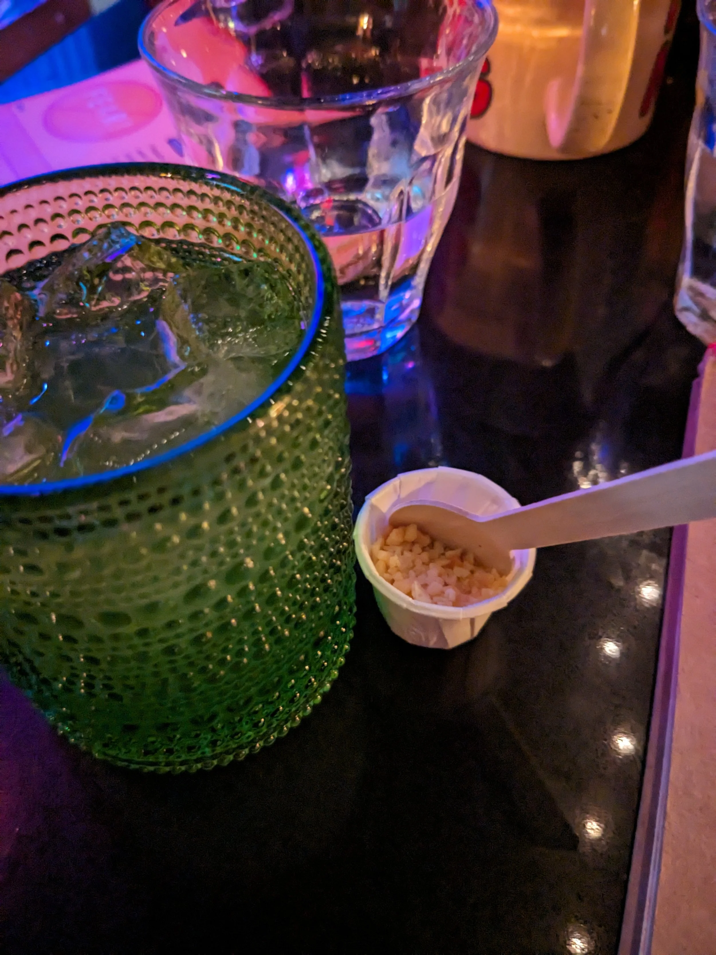 Close-up of two glasses with drinks, one with ice and a green textured glass, another clear glass with ice, a bowl of small snacks with a wooden spoon, and a brown container in the background on a dark table with purple lighting.