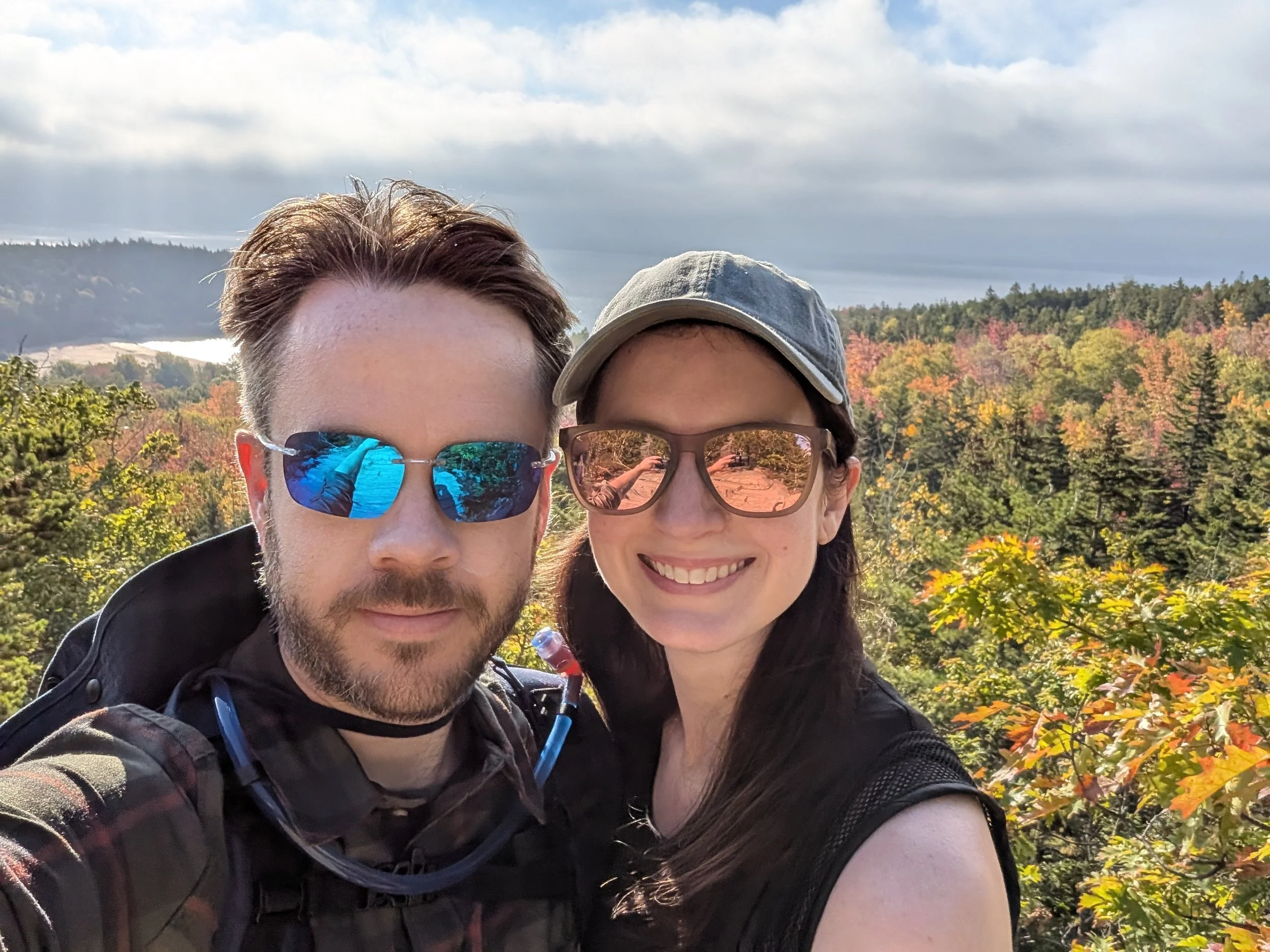Man and a woman smiling with trees of various colors and the ocean in the background  at Acadia National Park.