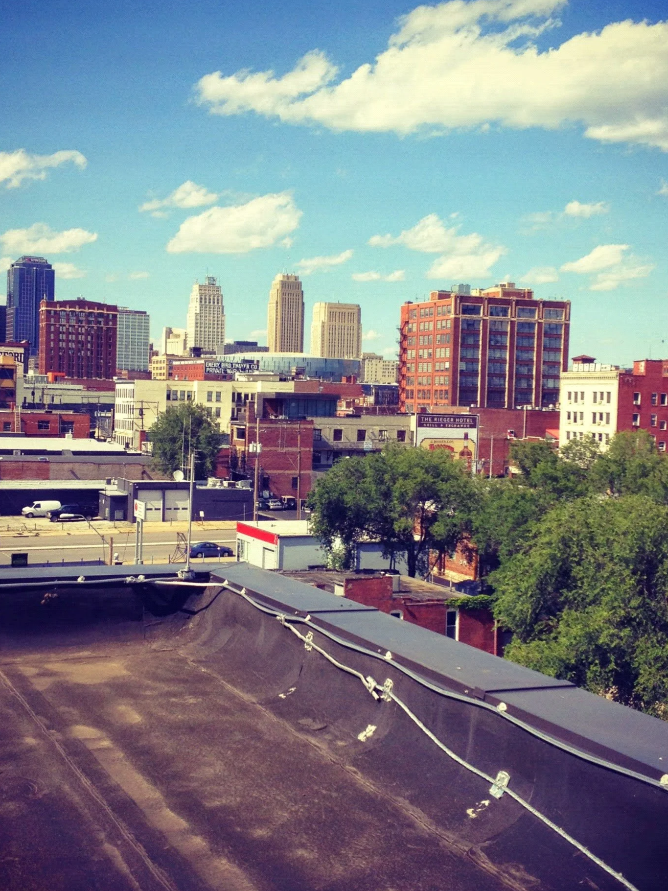 View of a city skyline with various tall buildings and skyscrapers under a blue sky with scattered clouds. The foreground shows rooftops and trees.