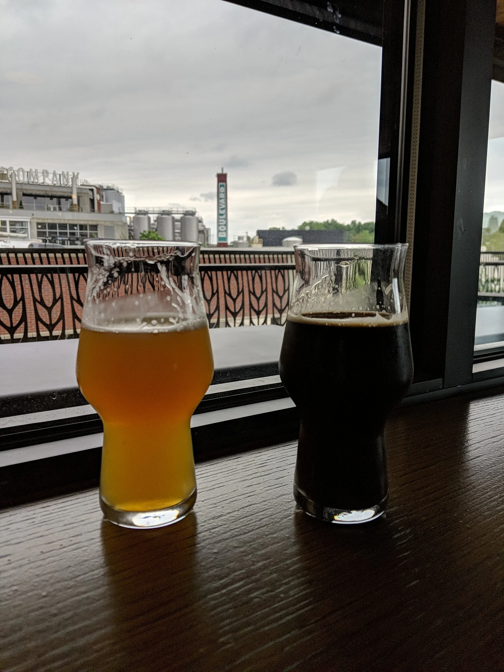 Two glasses of beer, one light and one dark, placed on a wooden table near a window with an industrial cityscape view.