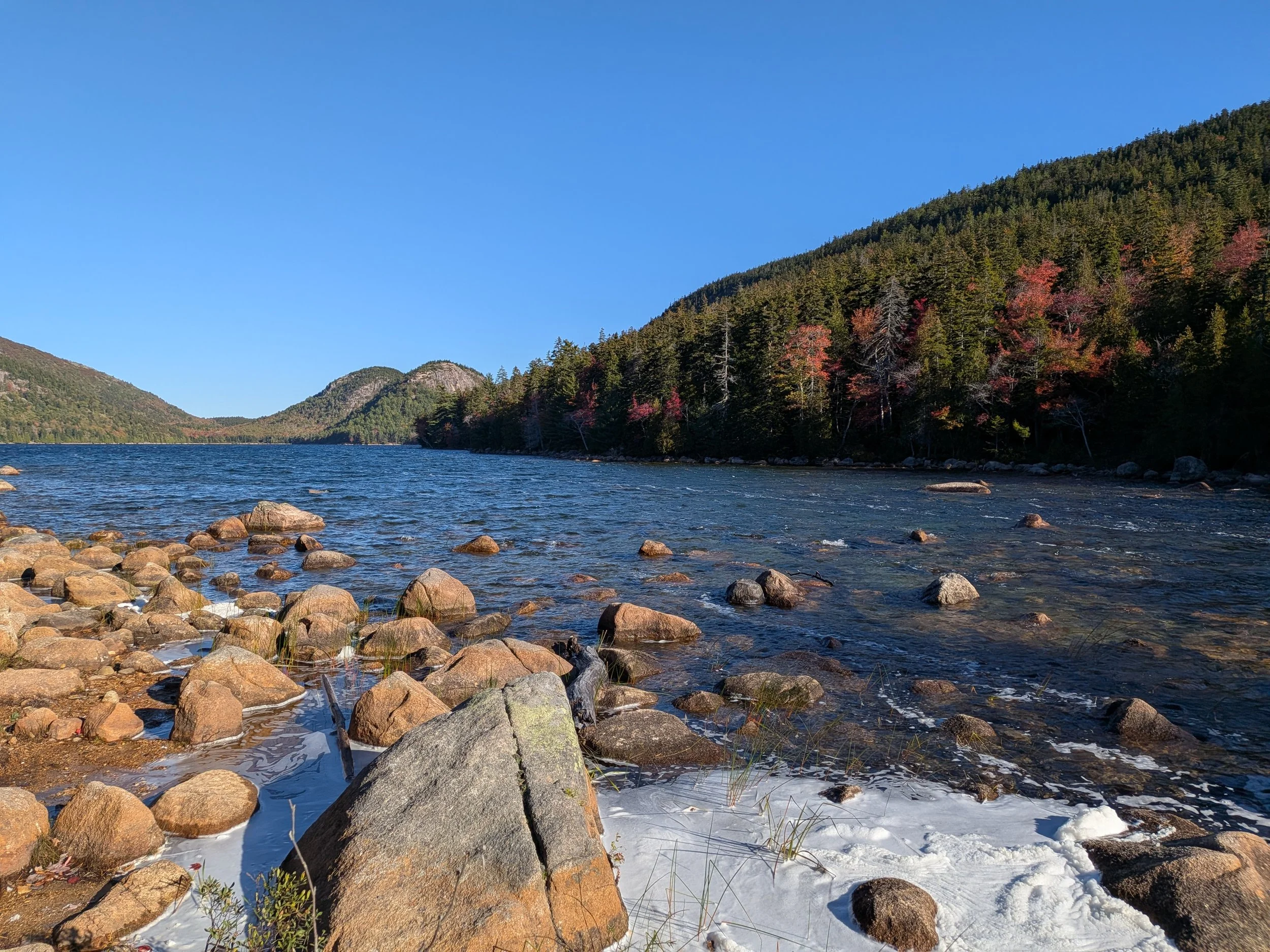 A lake with boulders and rocks in the foreground and trees and hills in the background.