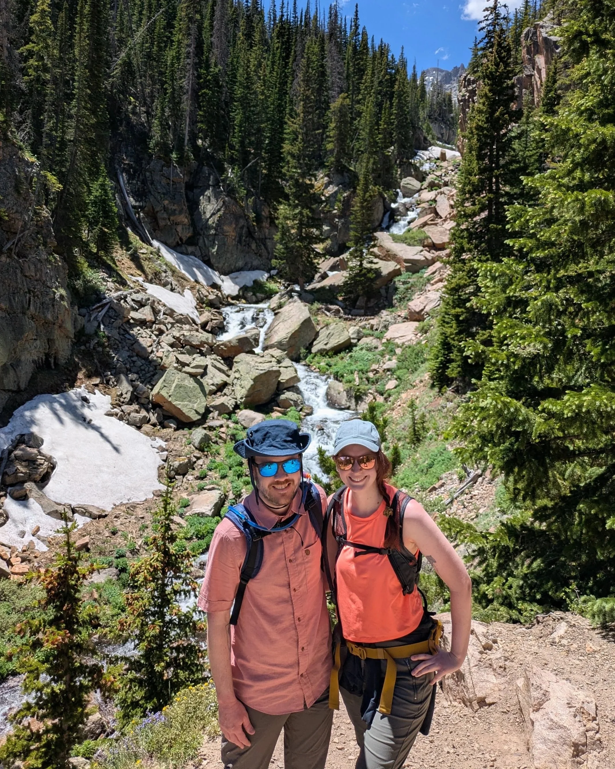A woman and a man standing in front of a waterfall among trees and a rocky cliff.