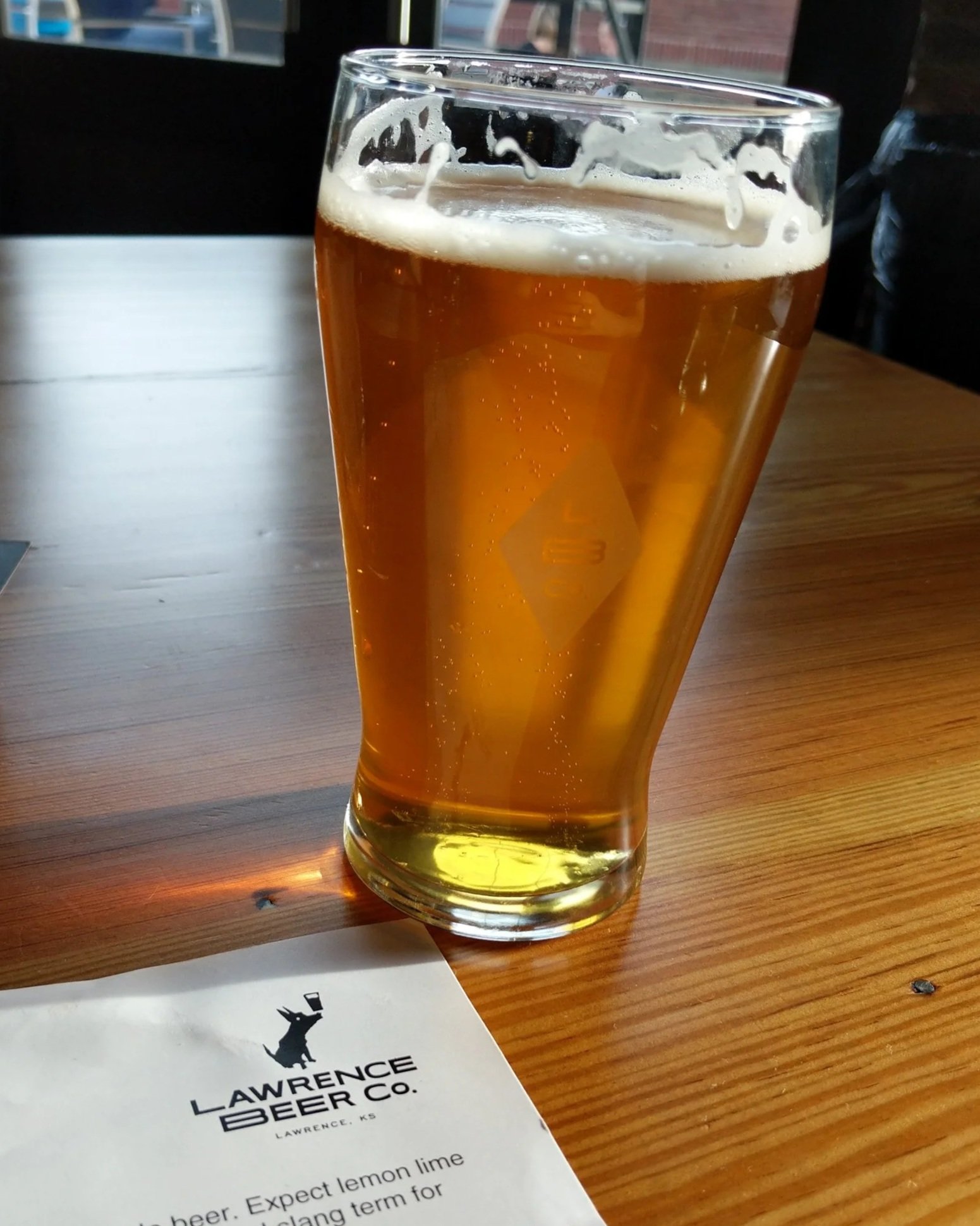 A pint of beer on a wooden table with a white menu that read "Lawrence Beer Co."